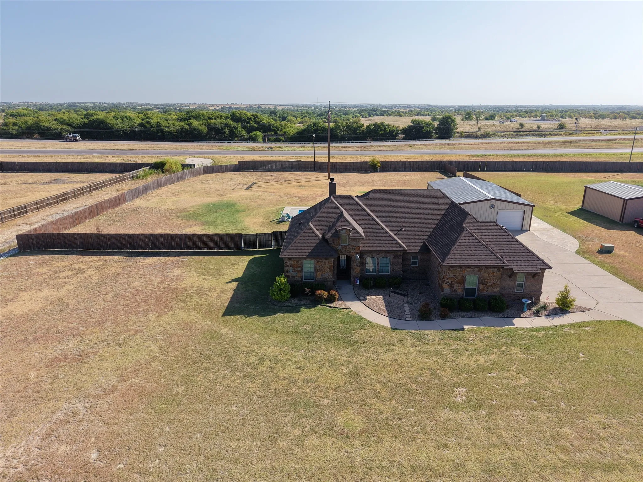 Brick residence situated on expansive acreage featuring a pitched roofline with dark shingle finish, multiple outbuildings including metal-frame structures, manicured lawn grounds, and established tree line bordering the property perimeter.