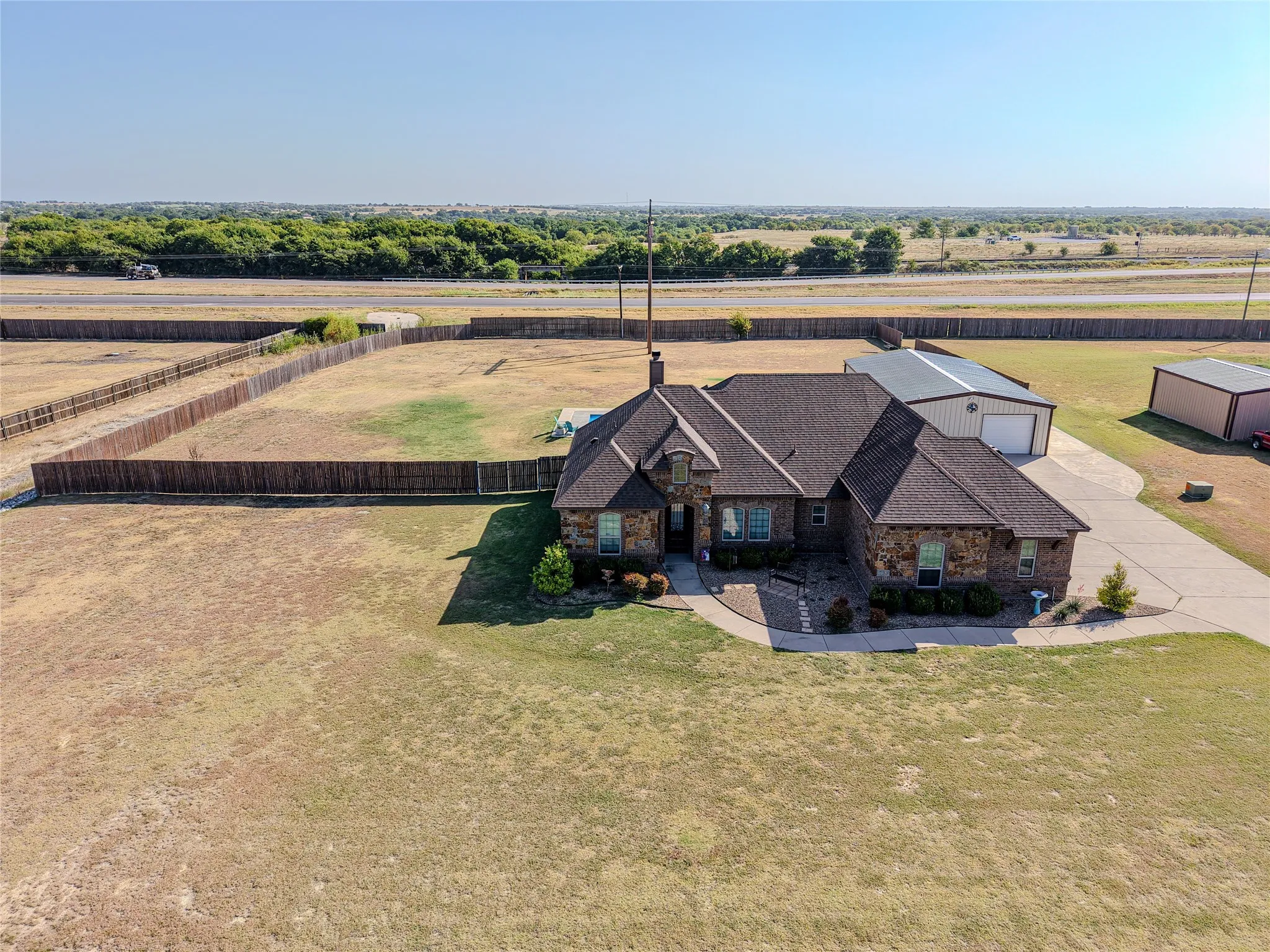 Stone and brick exterior with a dark shingle roof, concrete driveway, attached metal structure, and fenced perimeter