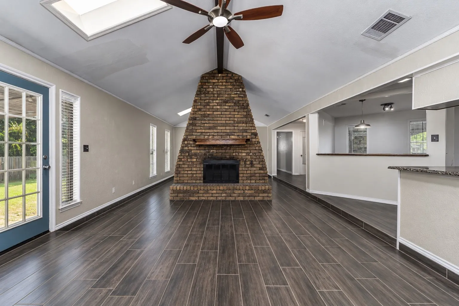 Vaulted ceiling with skylight and ceiling fan, brick fireplace with wood mantel, wood-look tile flooring, and a breakfast bar with stone-look countertop
