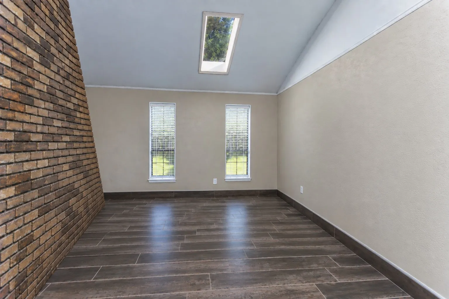 Vaulted ceiling with an integrated skylight, twin windows with blinds, brick-finish accent wall, and wood-finish tile flooring
