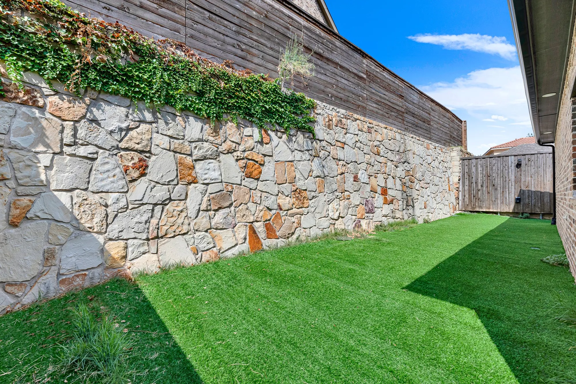 A towering native stone retaining wall crowned with climbing greenery forms a dramatic, private garden backdrop bathed in afternoon sun. Picture relaxing with a cool drink on warm evenings, enveloped by rustic textures and total backyard seclusion.