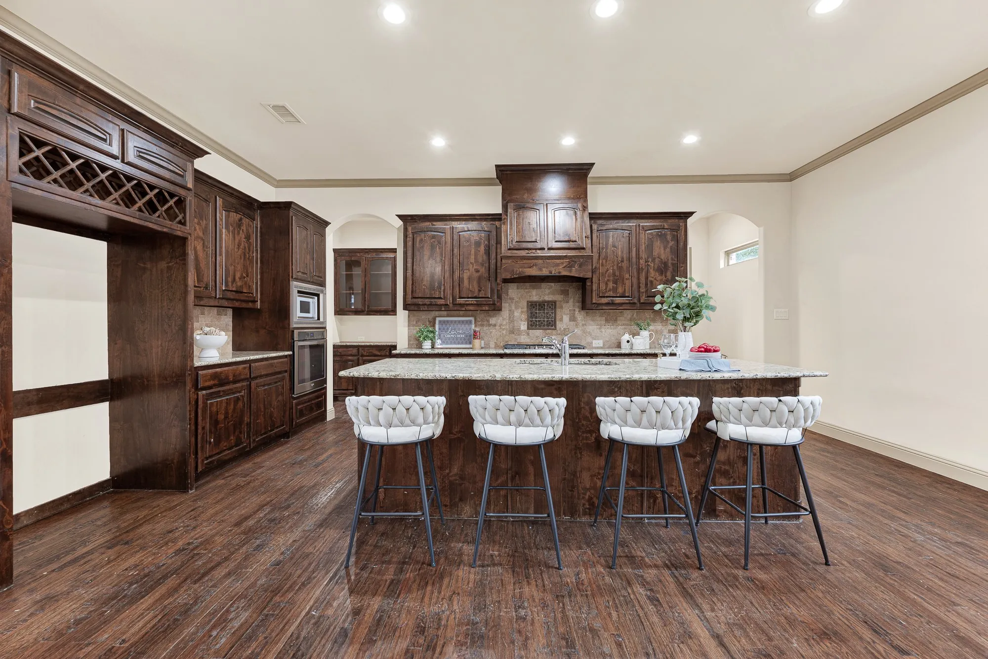 A striking carved wood range hood and rich espresso cabinetry anchor the back wall under crisp ceiling downlights. Imagine rolling out homemade pizza dough across the expansive granite island while family gathers around to help.