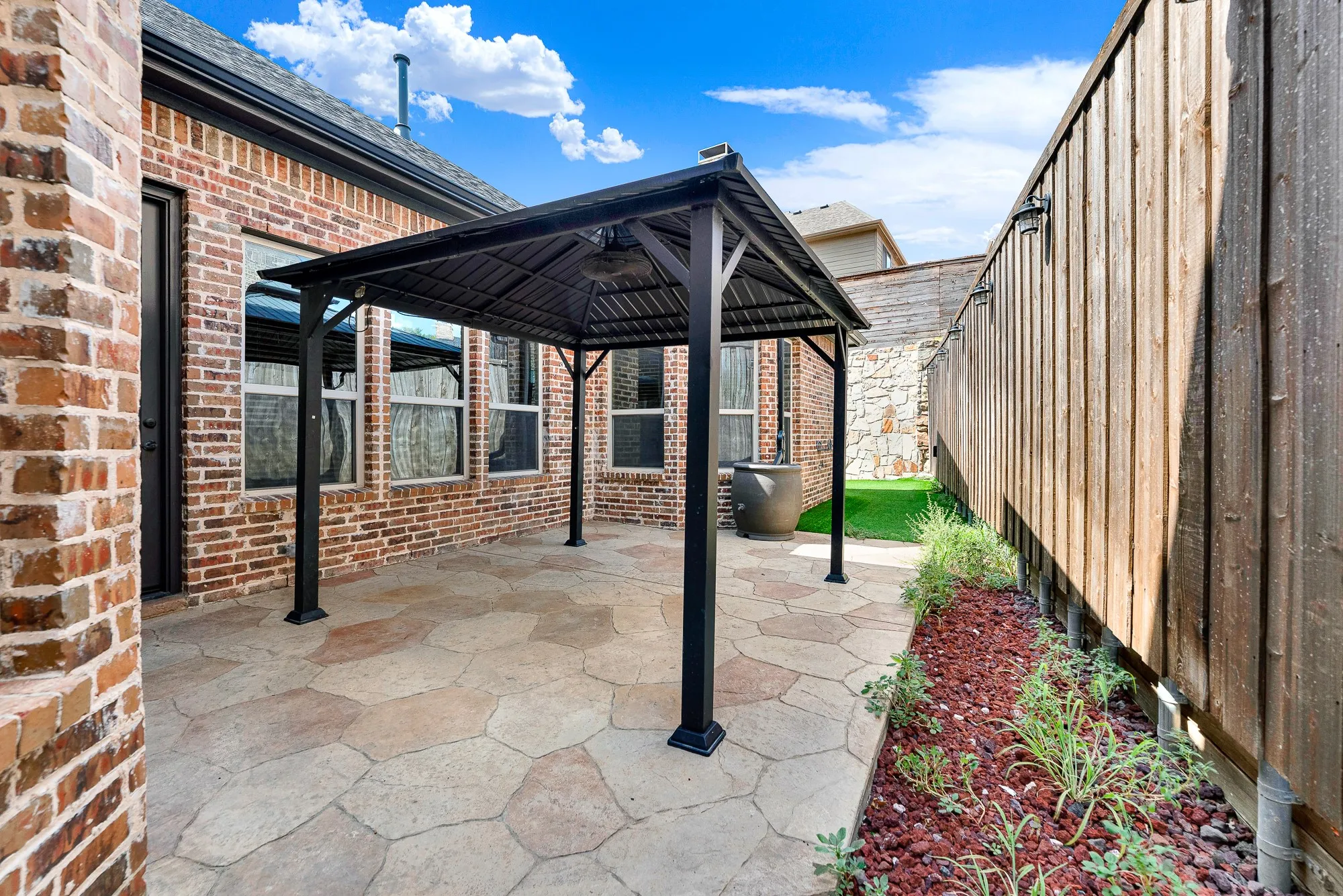 A stamped concrete courtyard extends beneath a dark metal-roofed pavilion, framed by privacy fencing and warm brick facades. It sets the scene for casual weekend barbecues, grilling burgers while friends gather comfortably in the open breeze.