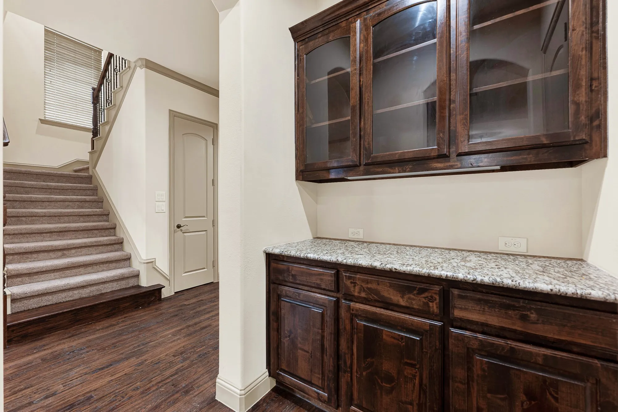 Glass-front upper cabinetry and dedicated granite serving surfaces tuck neatly beside the walk-in storage access and stairwell. It serves as an ideal staging station for mixing signature cocktails or decanting wine during dinner parties.