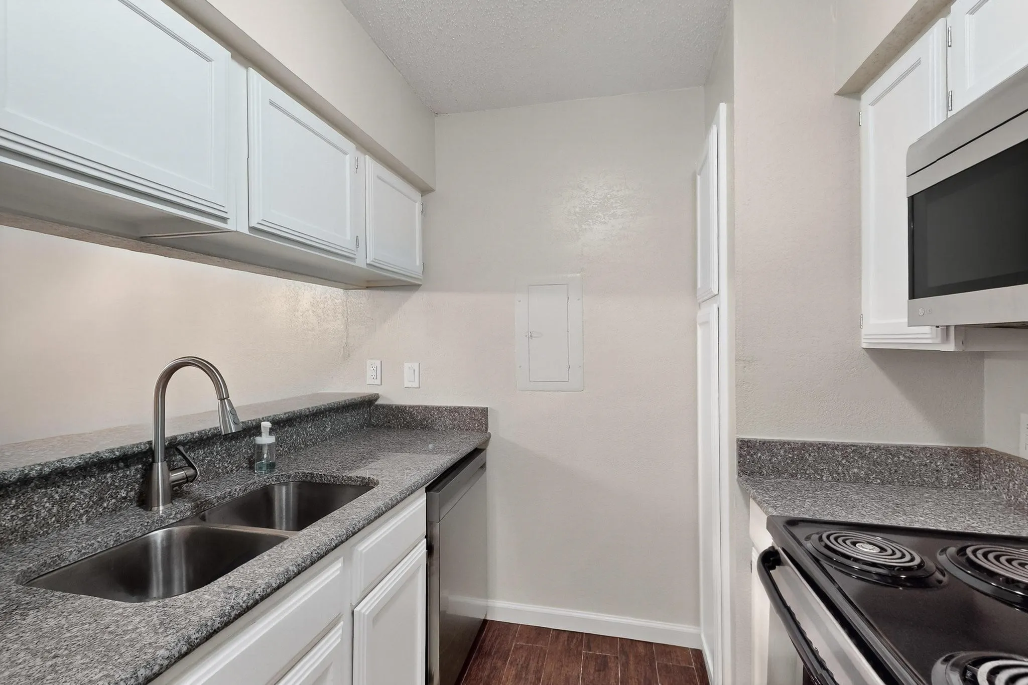 White upper and lower cabinetry, dual basin stainless steel sink with gooseneck faucet, dark speckled countertops, wood-finish flooring, and a stainless steel over-the-range microwave