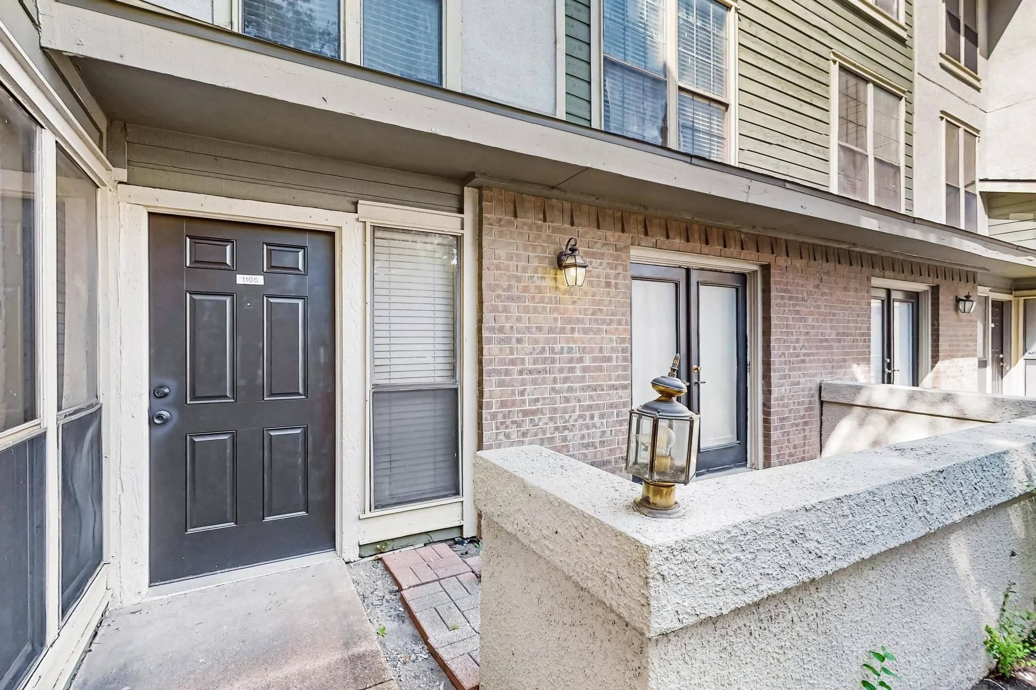 Brick exterior with concrete patio, black paneled entry door, adjacent window, and outdoor lighting fixtures