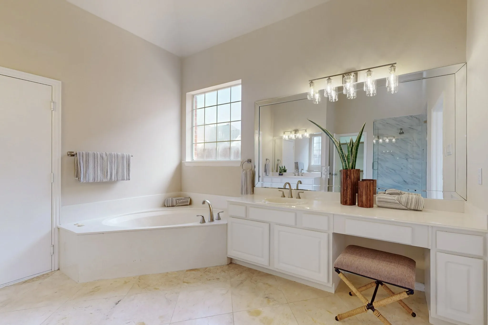 Bright primary bathroom with soaking tub, large white vanity, oversized mirror, stone tile flooring, modern lighting, and a seated dressing area