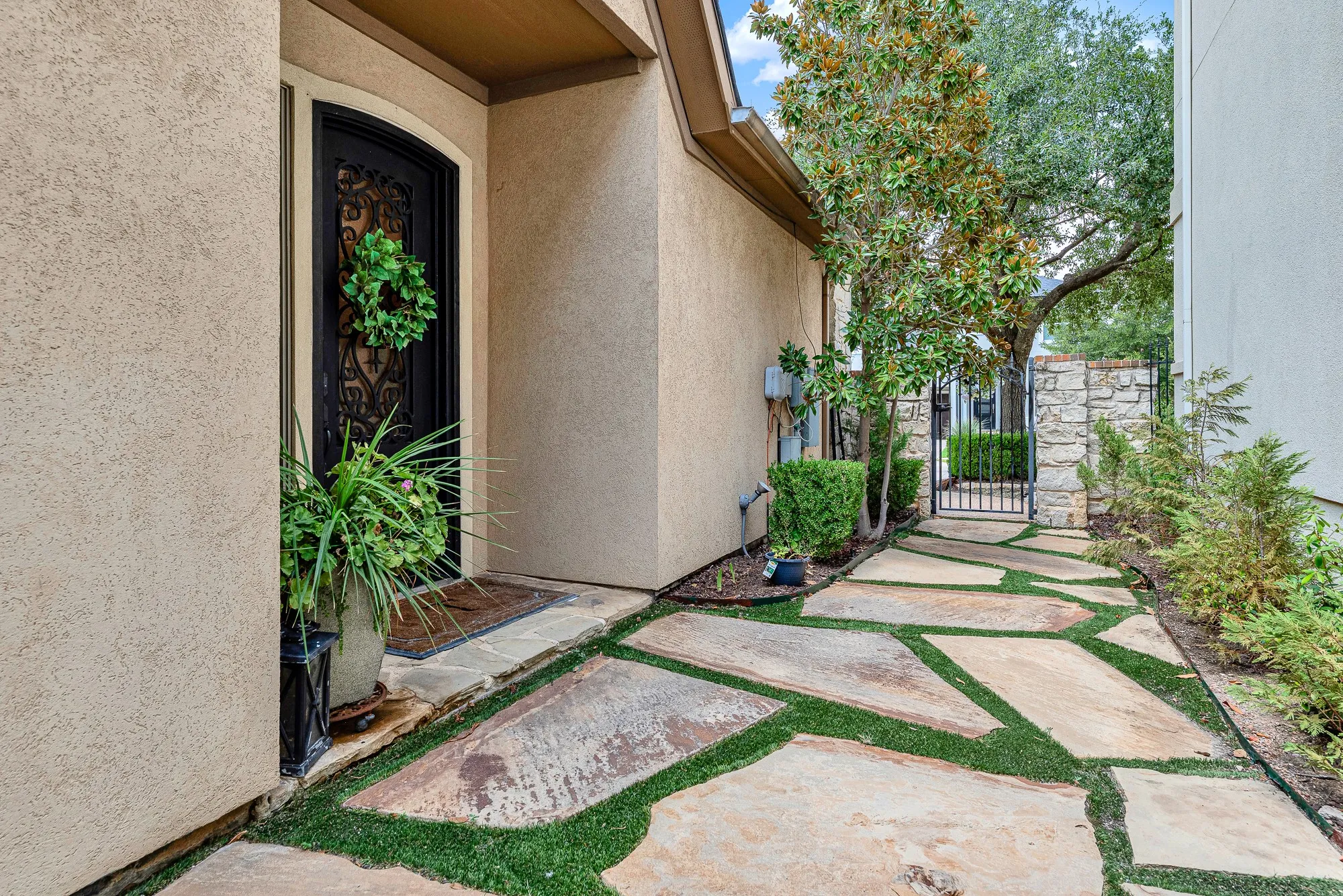 A wrought-iron gate frames the walkway while tall alcove walls cast deep, cool shadows across the pathway. Picture welcoming close friends through this secret garden path as twilight sets in for an intimate evening dinner.