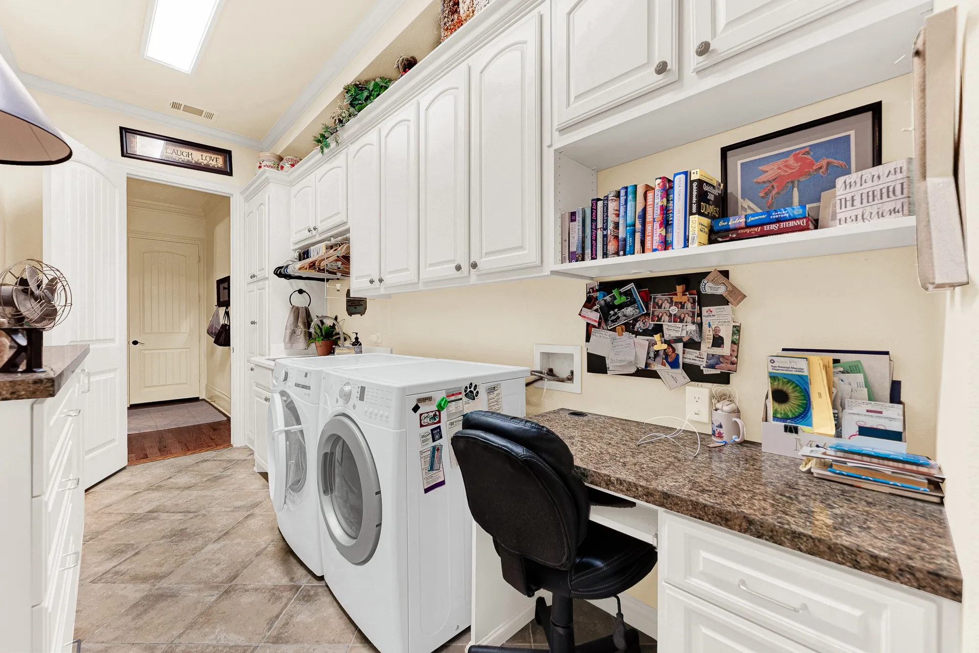 Bright white upper cabinetry and a long built-in desk stretch across durable tile flooring, illuminated by clean overhead lighting. Sit down to organize weekly schedules or manage family affairs in this quiet, efficient workspace.
