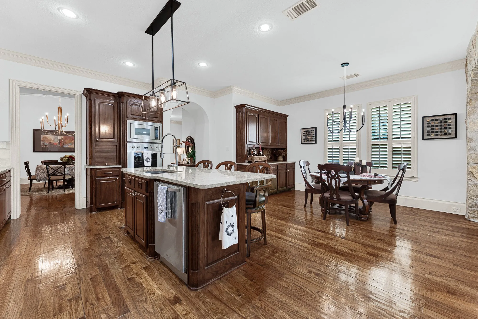 Shuttered windows flood the casual dining space with gentle sunlight beside a custom built-in beverage station. Enjoy relaxing Sunday morning breakfasts bathed in natural light before starting your day. Built-in stainless steel appliances blend into rich wood cabinetry under glowing task lighting. Imagine prepping a favorite family recipe with every essential tool conveniently within arm’s reach.