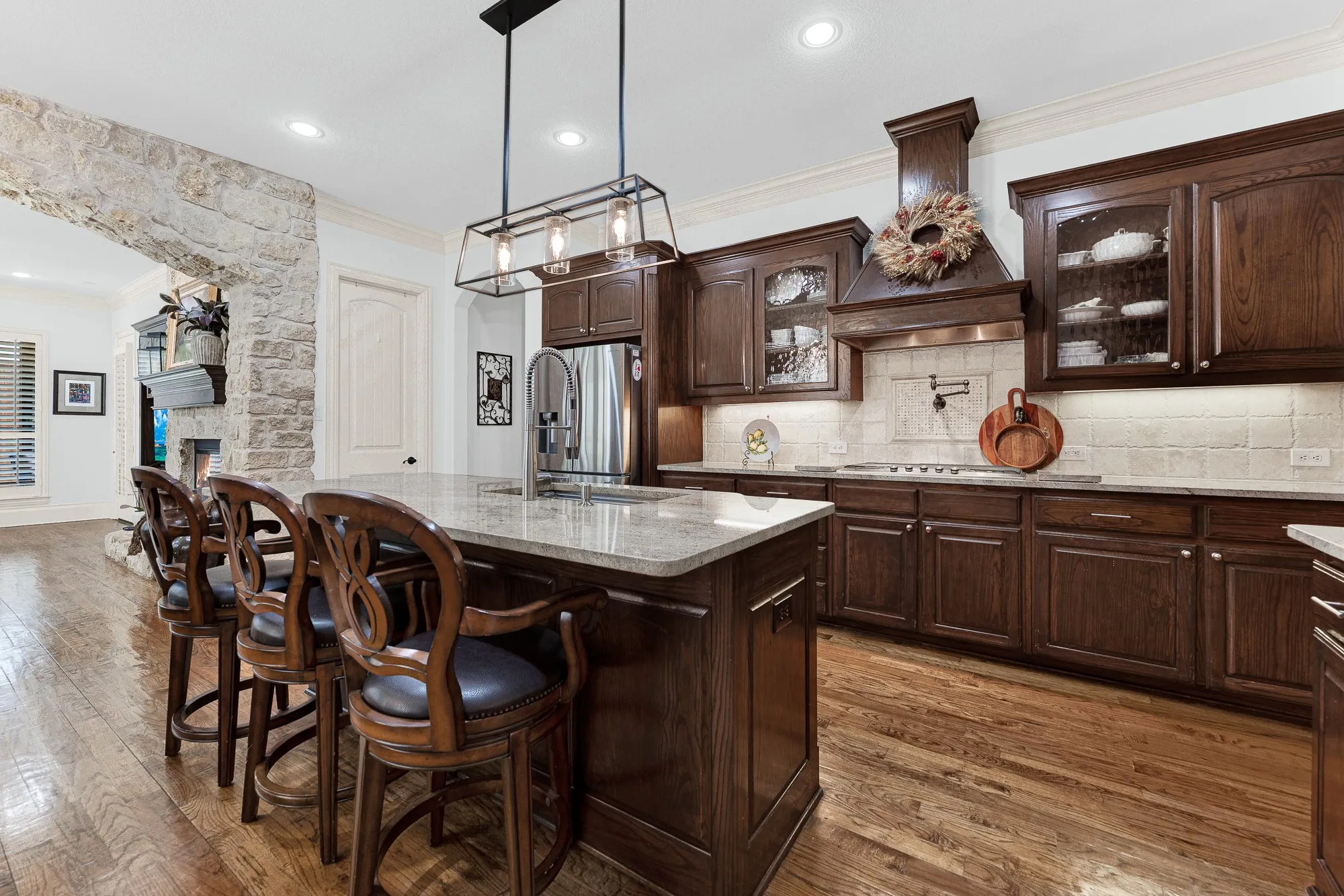 Polished granite countertops reflect linear pendant lights, surrounded by deep-toned cabinetry and detailed molding. Visualize plating gourmet weekend hors d'oeuvres while guests gather comfortably around the central island.