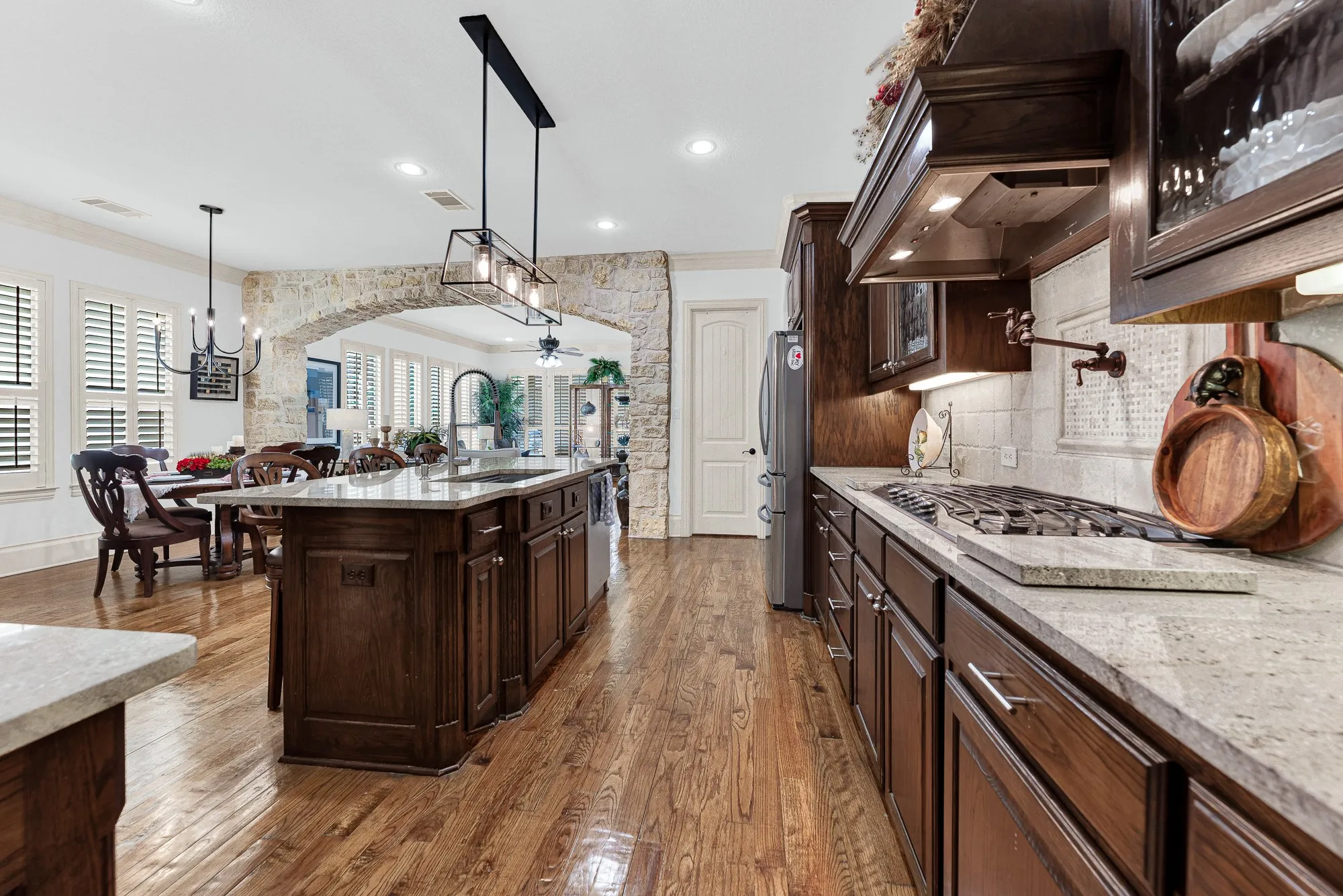 A majestic stone archway anchors the space, bridging culinary prep areas with adjacent social nooks under soft recessed lighting. Picture sipping espresso at the island bar while chatting with family lounging nearby. A textured tile backsplash and decorative vent hood frame the gas cooktop with pot filler, highlighted by warm under-cabinet lights. Savor the mouthwatering aromas of a simmering dinner filling the air as evening approaches.