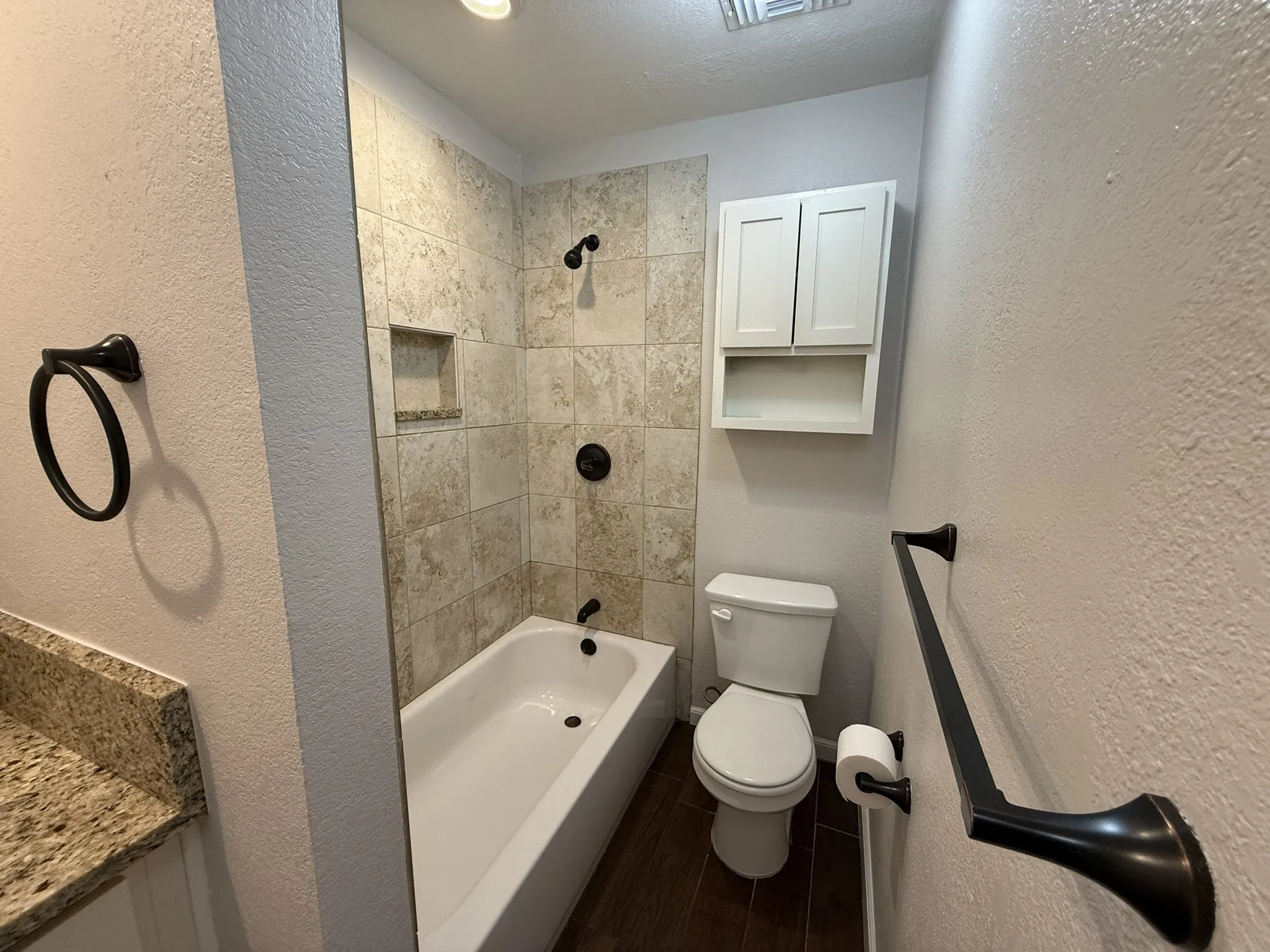 Tiled bath and shower combination with a recessed niche, dark bronze fixtures, wood-finish flooring, and a stone-finish countertop