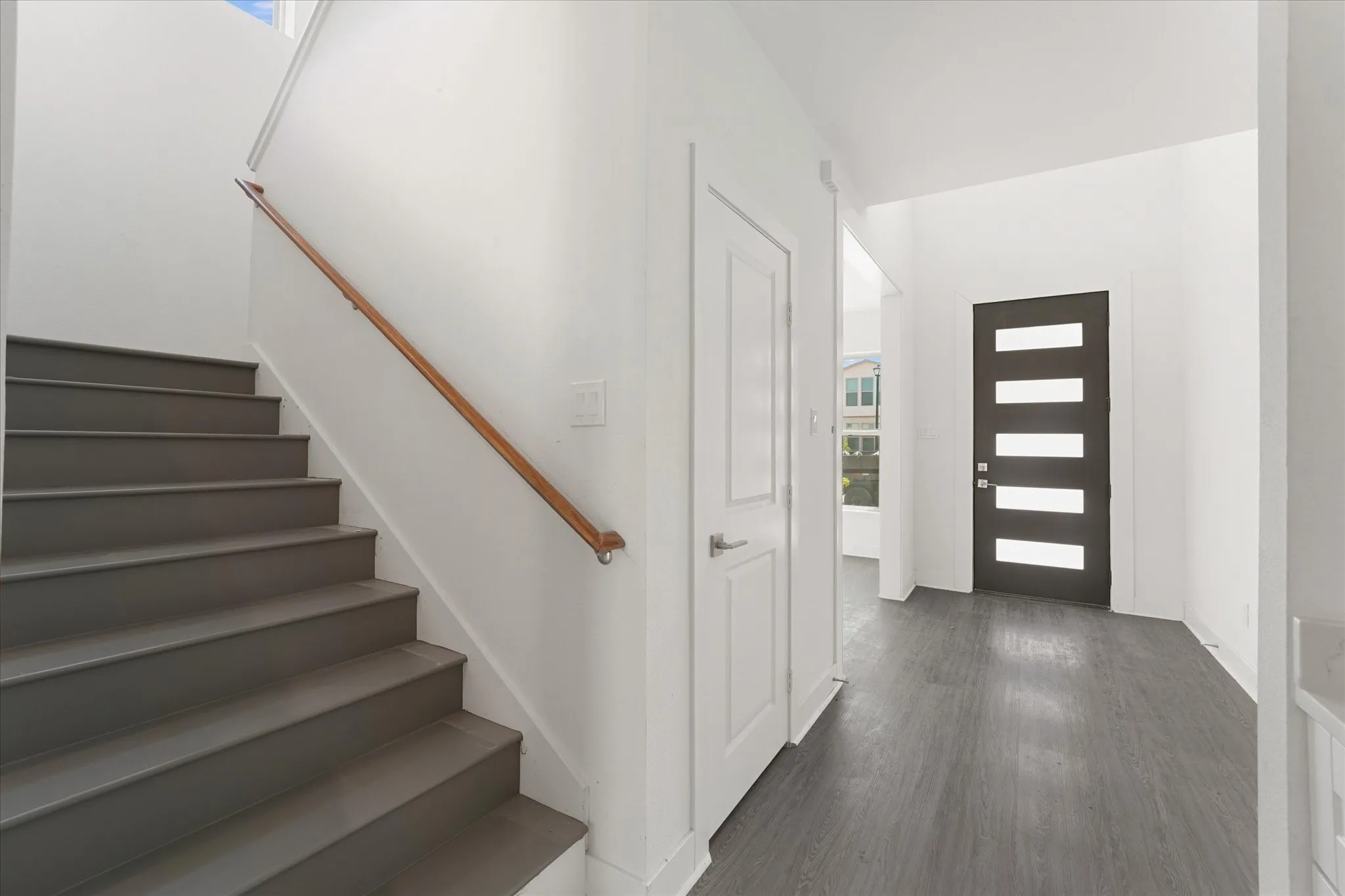 Wood-finish flooring throughout the entry, modern dark front door with horizontal glass panels, and a painted staircase with a wood handrail