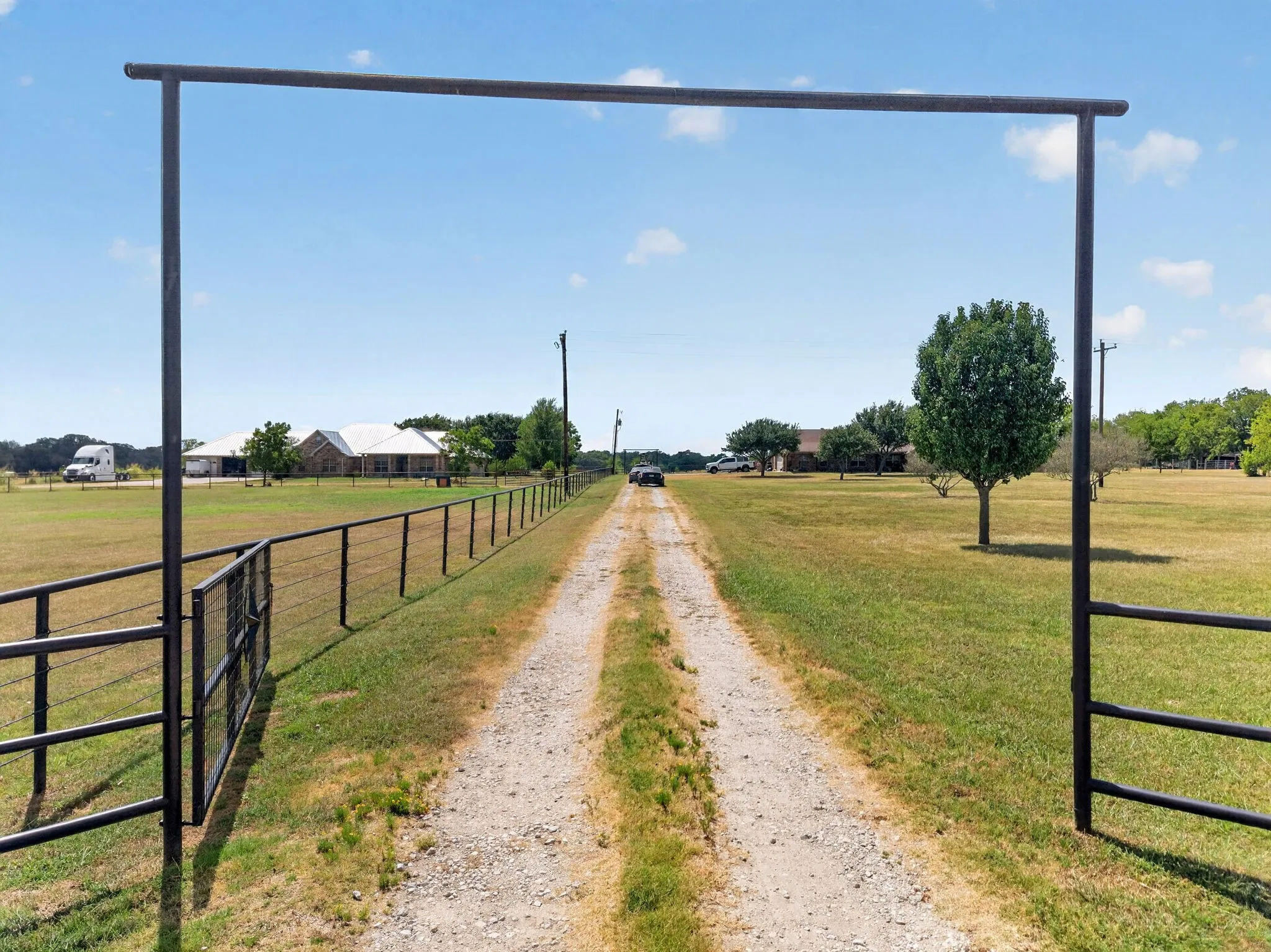 Gravel driveway leading to the property, featuring a metal entry gate, perimeter fencing, and expansive green spaces