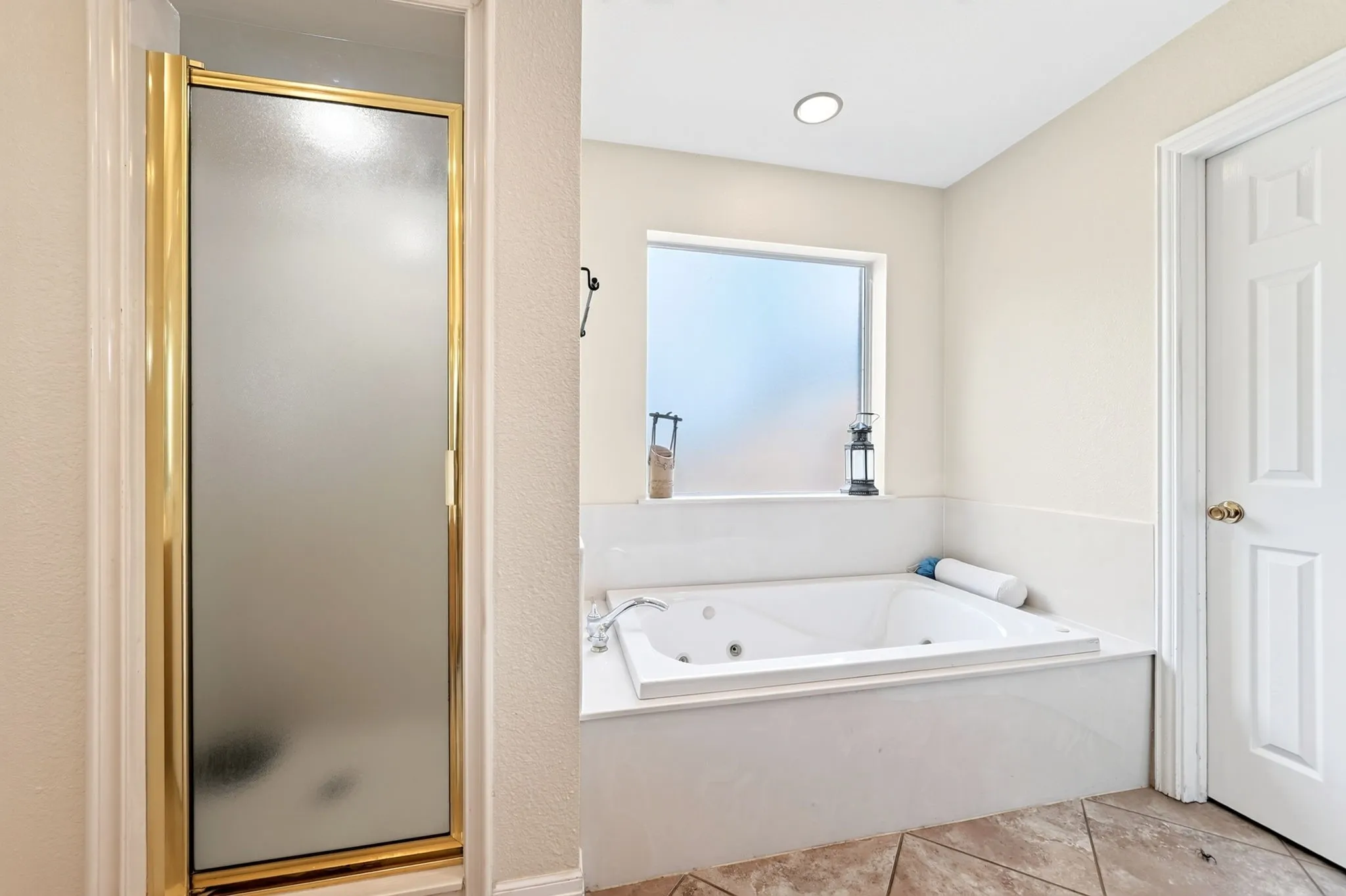 Recessed lighting, large soaking tub, tiled tub surround, frosted window, and gold-finish shower enclosure