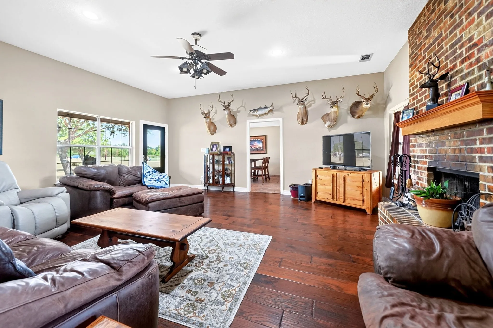 Warm living area featuring rich wood-finish flooring, a brick fireplace with a wood mantel, and expansive windows