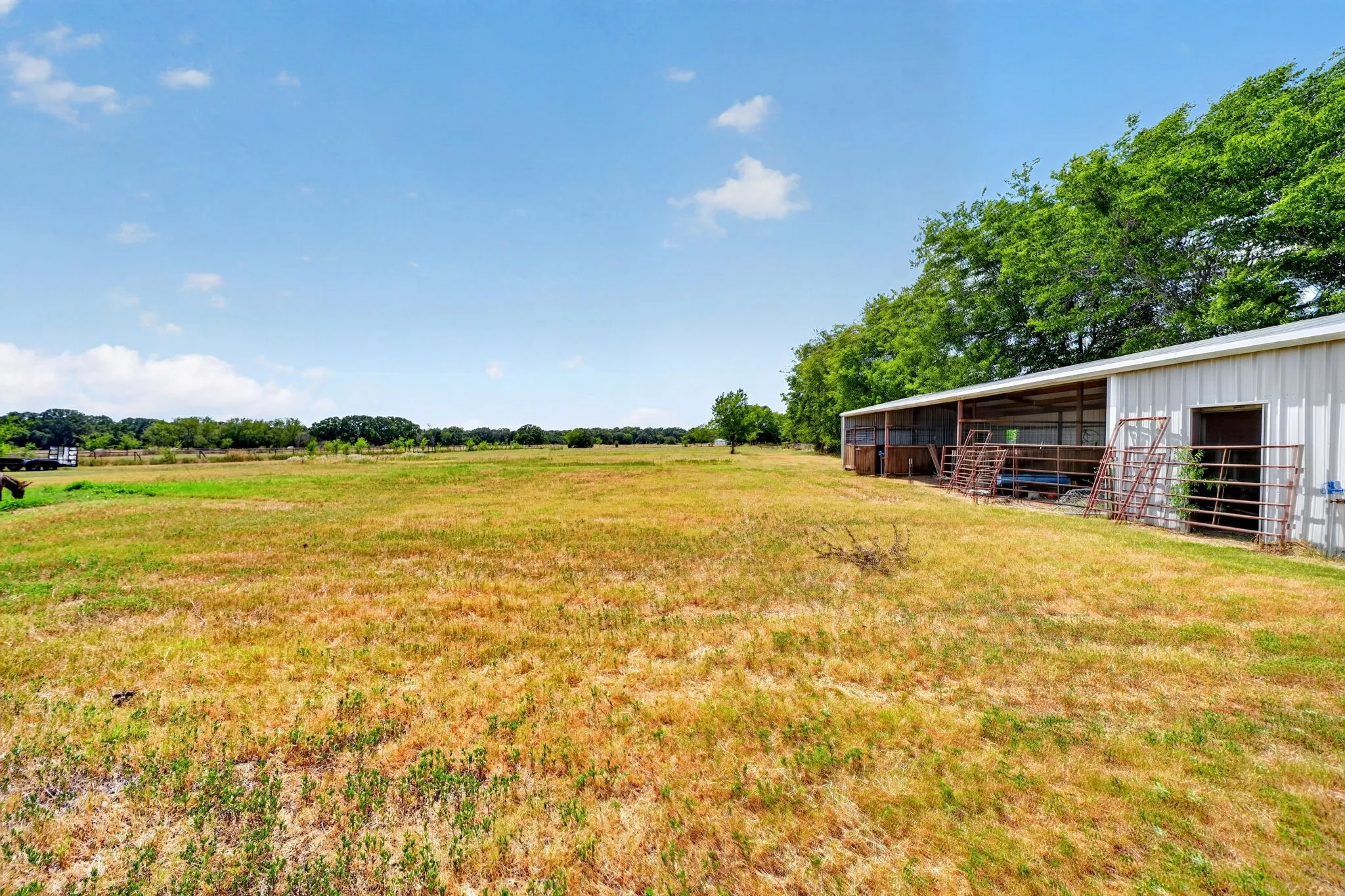 Expansive open field with a large metal-sided outbuilding featuring multiple covered stalls and a fenced enclosure