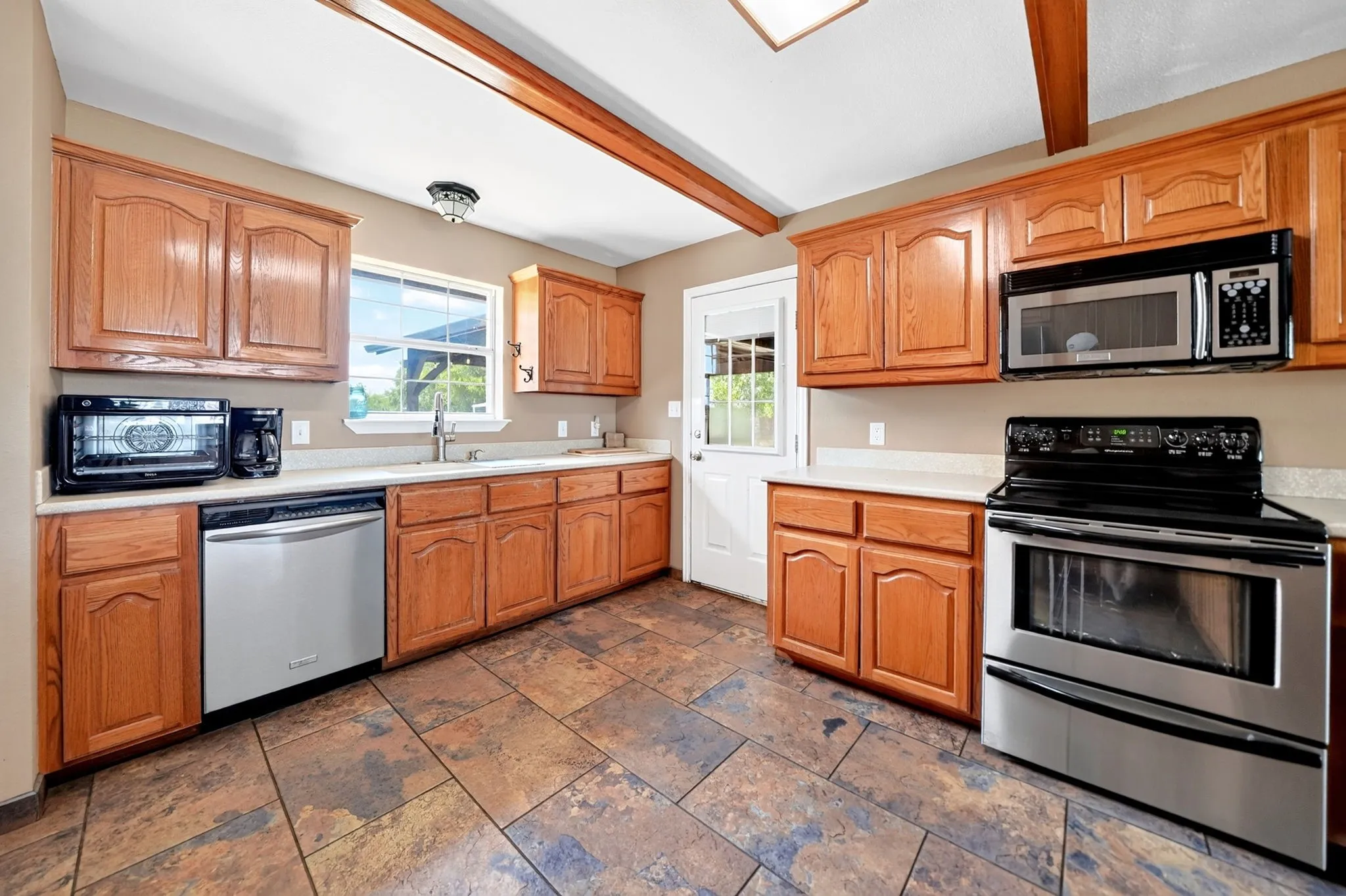 Kitchen featuring wood cabinetry, stainless steel appliances, light-toned countertops, and exposed ceiling beams
