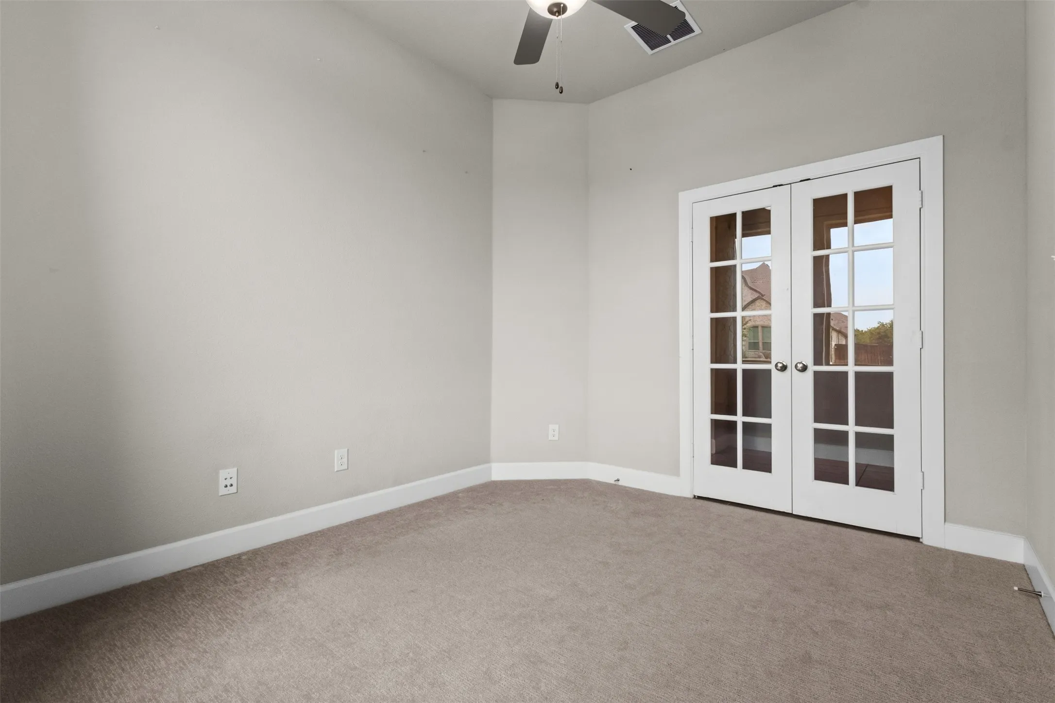 Bright, neutral-toned bedroom featuring neutral carpeting, white-trimmed French doors with multiple glass panes, ceiling fan, and multiple wall outlets for convenient placement of furnishings and electronics