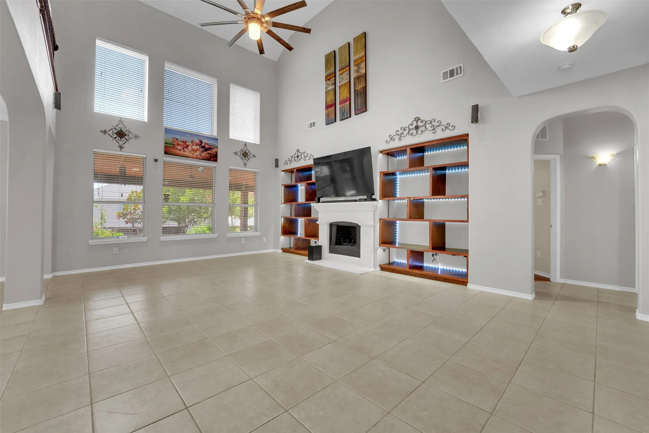 Vaulted ceiling with ceiling fan, tile flooring, built-in shelving with accent lighting, fireplace with mantel, and multiple windows