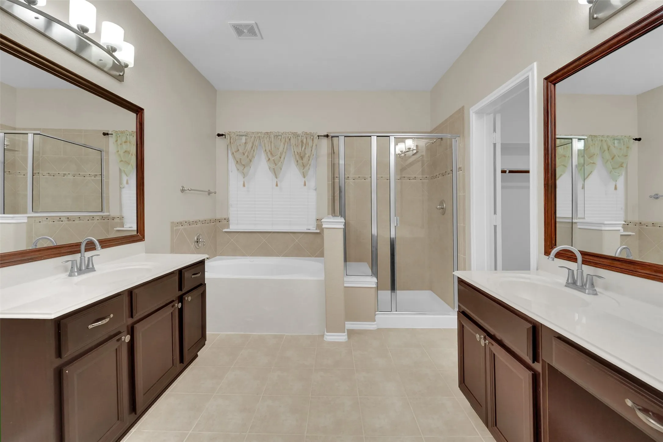 Dual vanity with integrated sinks, framed mirrors, a built-in soaking tub, a tiled walk-in shower with glass enclosure, and neutral tile flooring