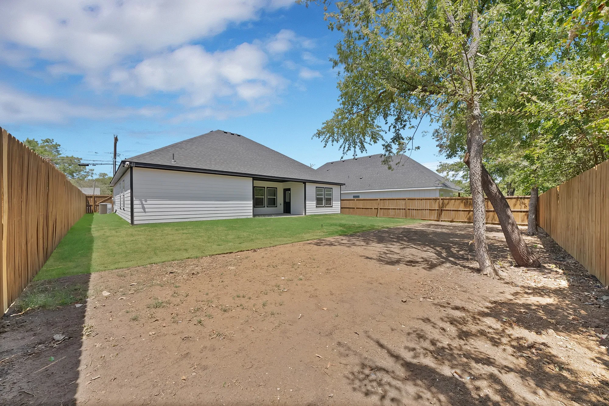 Expansive backyard featuring a new wood privacy fence, partial grass lawn, mature trees, and a covered patio