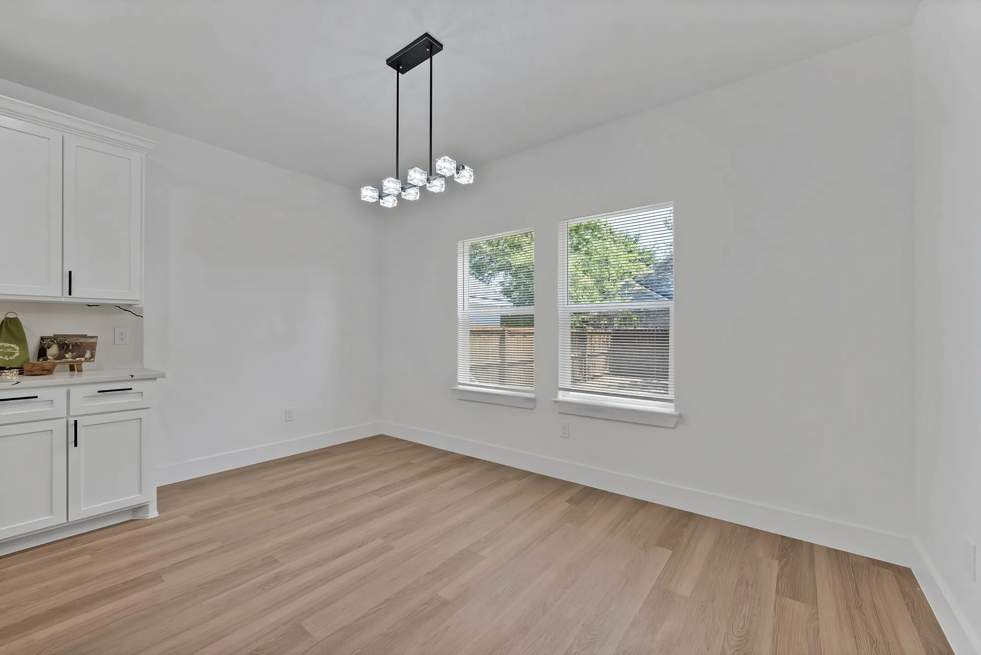 White cabinetry with matte black hardware, light wood-finish flooring, two large windows, white walls, and a contemporary linear chandelier
