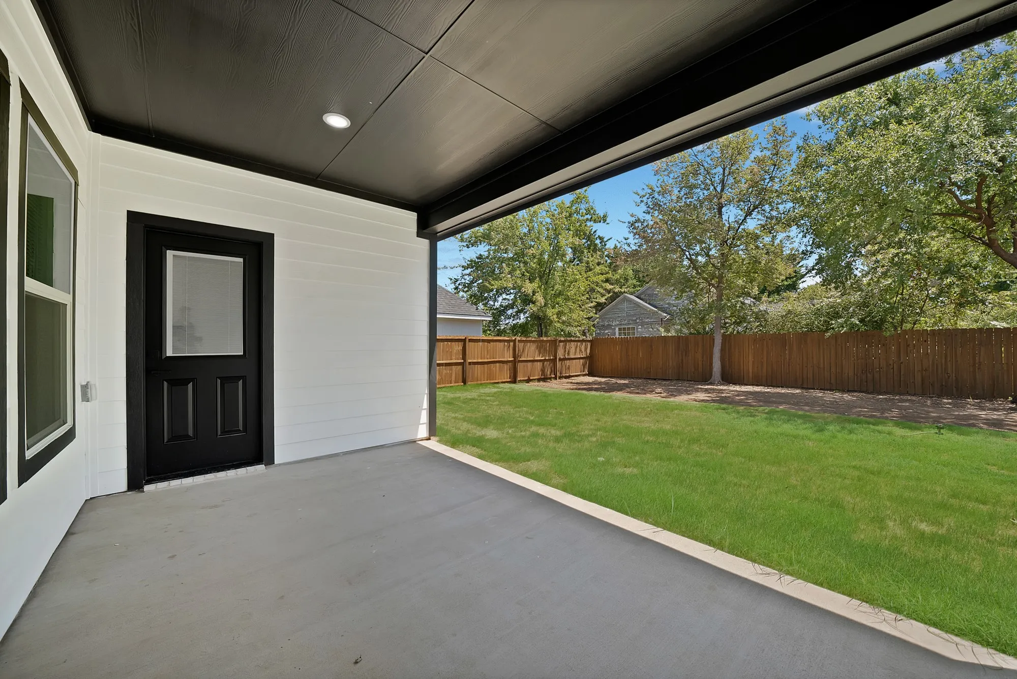 Covered concrete patio with recessed lighting, offering access to an expansive fenced yard featuring a manicured lawn and mature trees