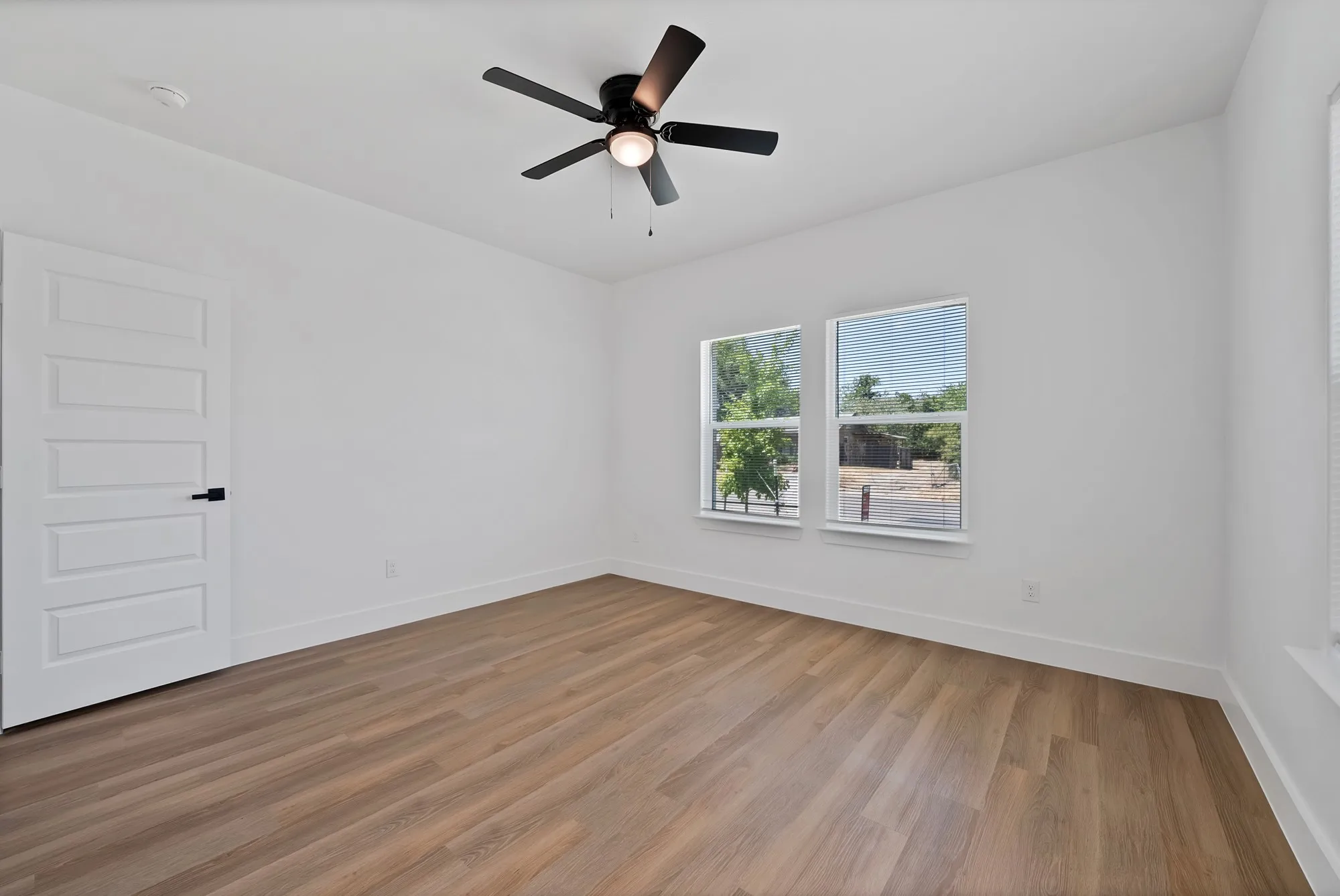 Wood-finish flooring, white walls, two windows, ceiling fan, and a paneled door with matte black hardware