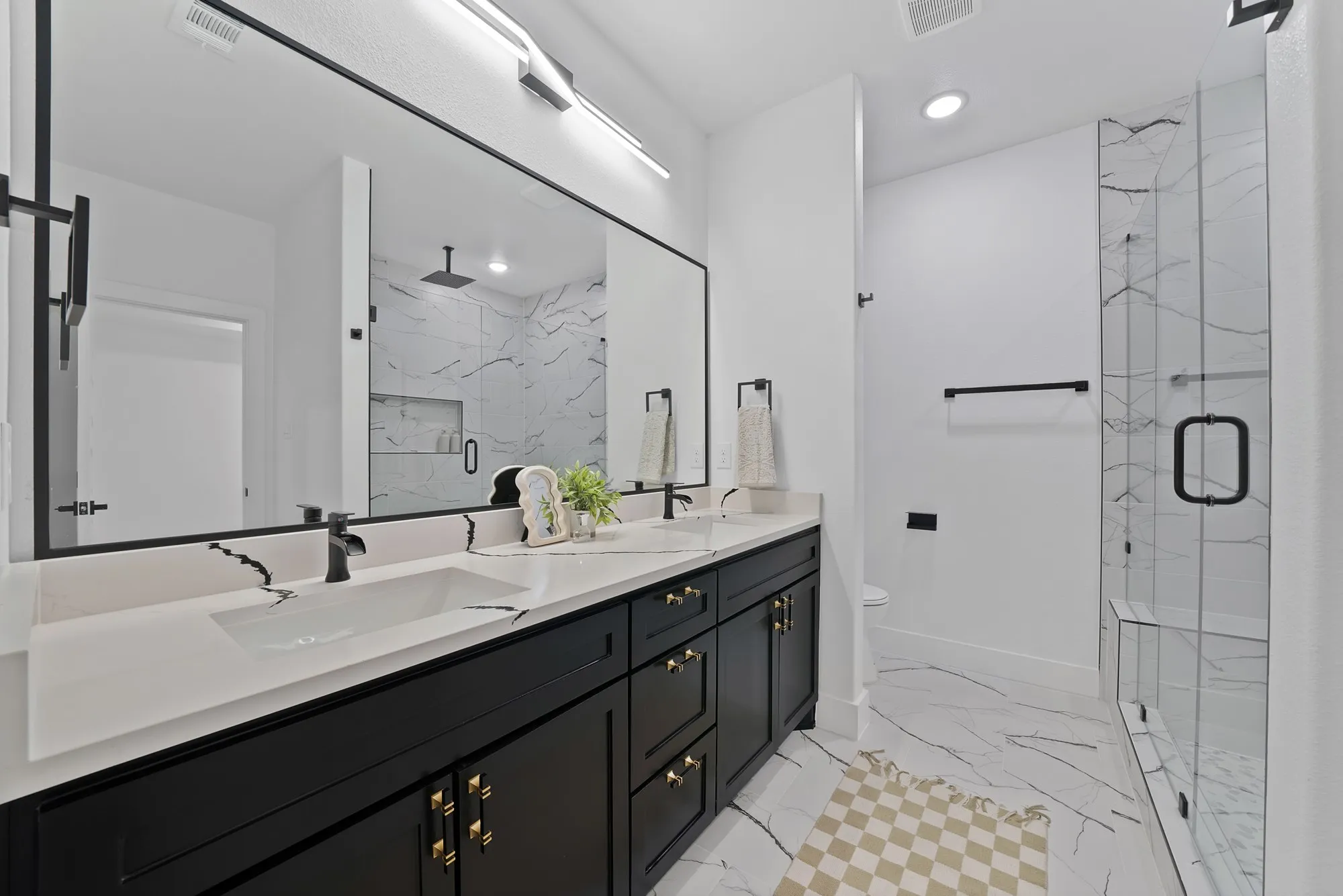 Dual vanity with undermount sinks, quartz countertops, black cabinetry with gold-tone hardware, and a frameless glass shower with a built-in bench