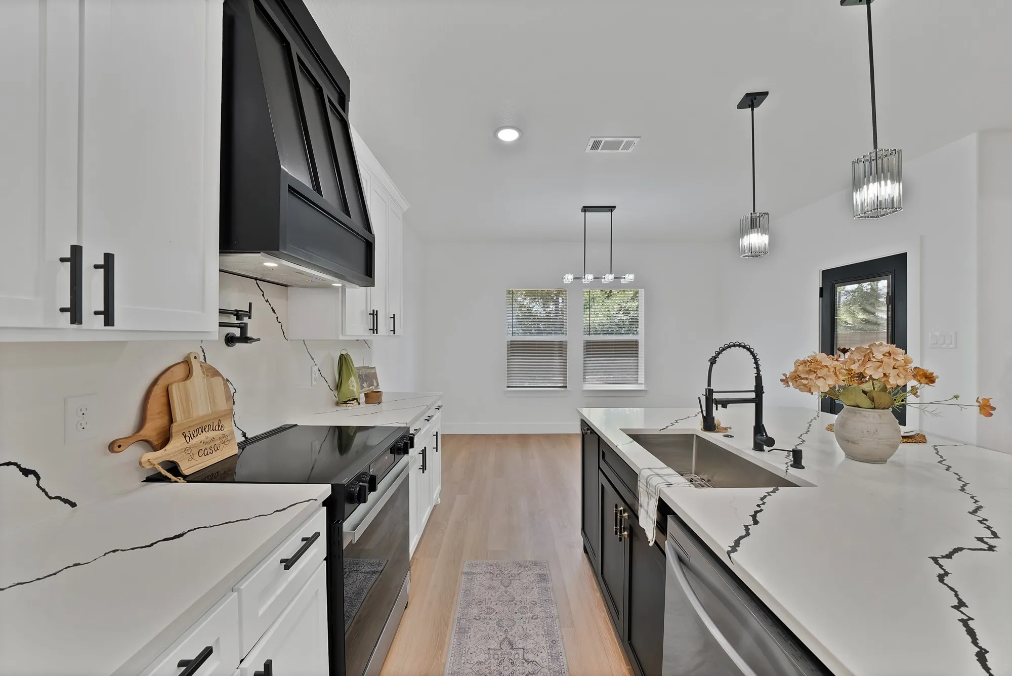 White cabinetry with matte black hardware, black range hood, light-toned wood-finish flooring, white countertops with prominent black veining, and a kitchen island with a built-in sink and pendant lighting