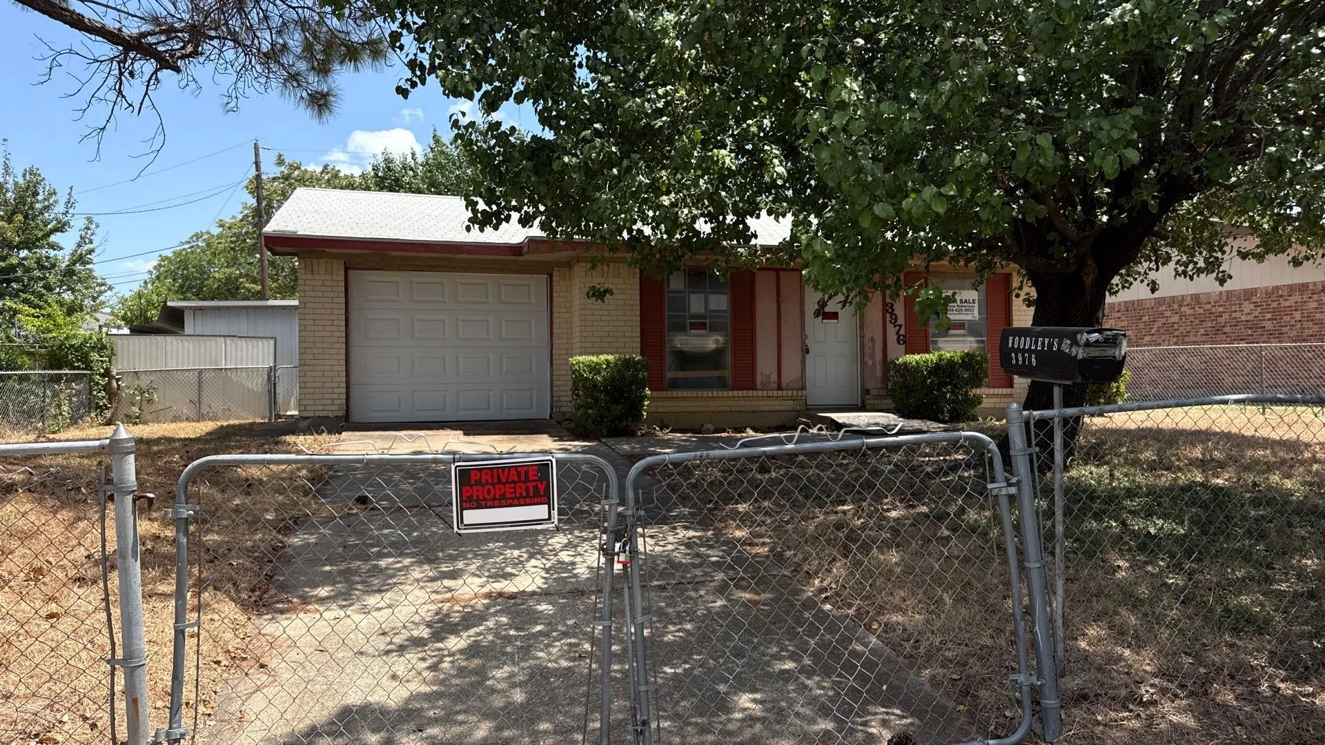 Chain-link fence with a double gate, concrete driveway, attached garage, brick exterior, and mature trees