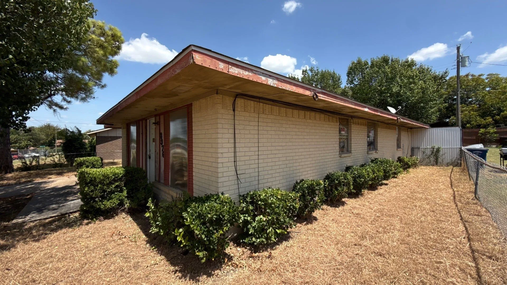 Light-colored brick exterior featuring a red soffit and fascia, with a row of manicured shrubs along the foundation
