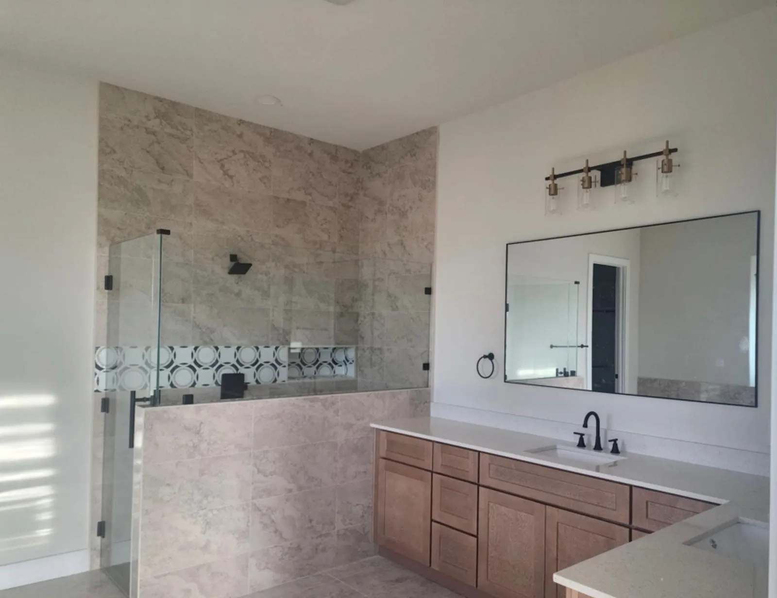 Frameless glass shower enclosure with decorative tile inlay, large format wall tiling, wood-finish vanity cabinetry, dual vanity sinks, and a wide framed wall mirror
