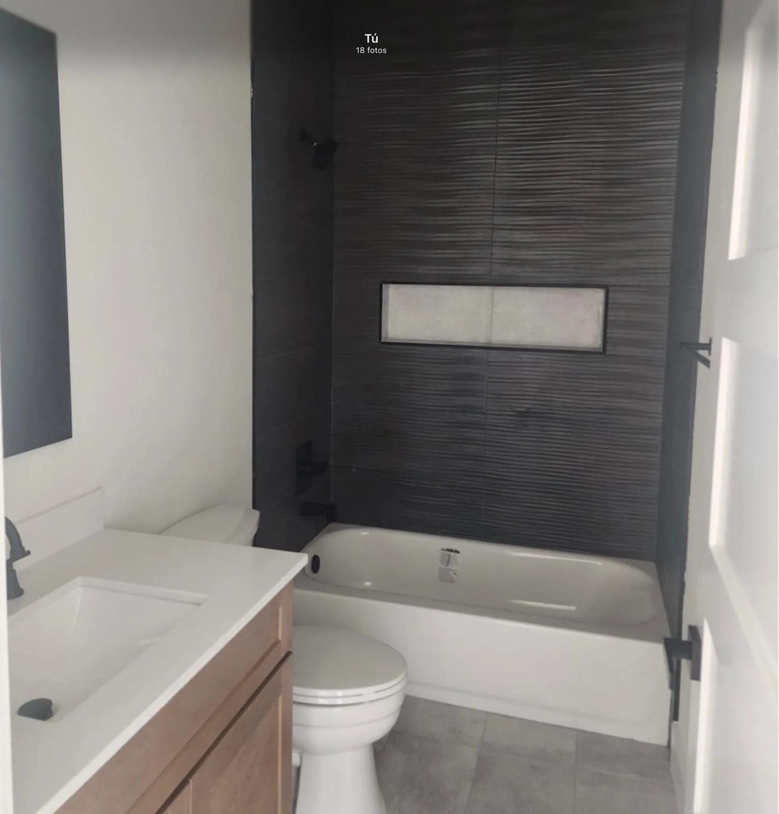 Bathroom featuring a wood-finish vanity with an integrated sink, a white bathtub, dark textured tile shower surround, and a recessed shower niche
