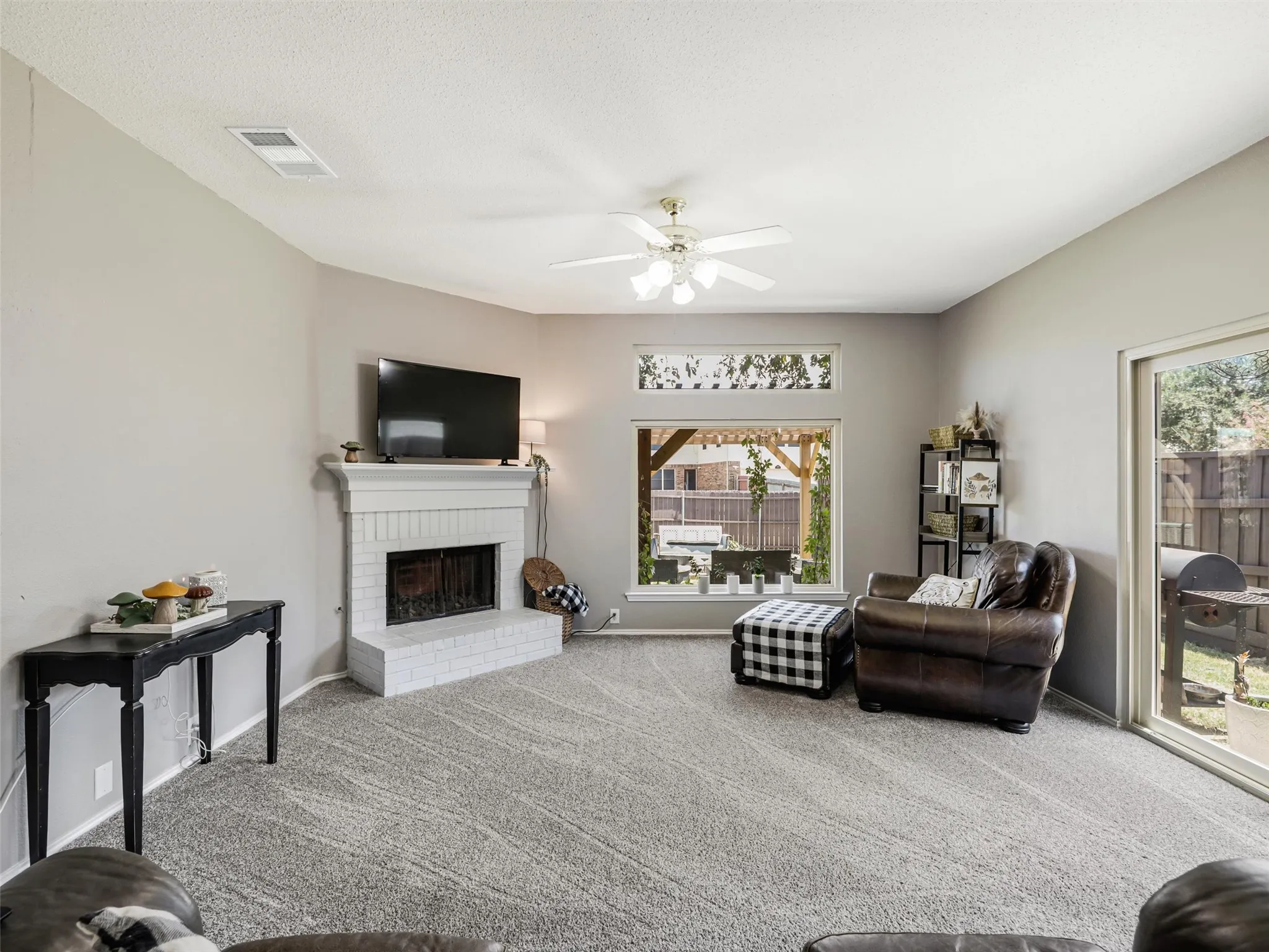 Carpeted living area featuring a white painted brick fireplace, ceiling fan, and large windows with transom lighting