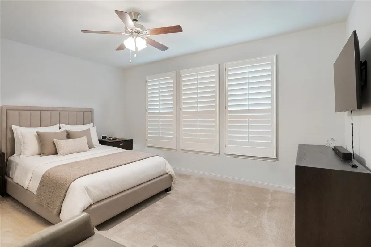 Plush carpeting, three windows with plantation shutters, ceiling fan with integrated lighting, and a dark wood-finish nightstand
