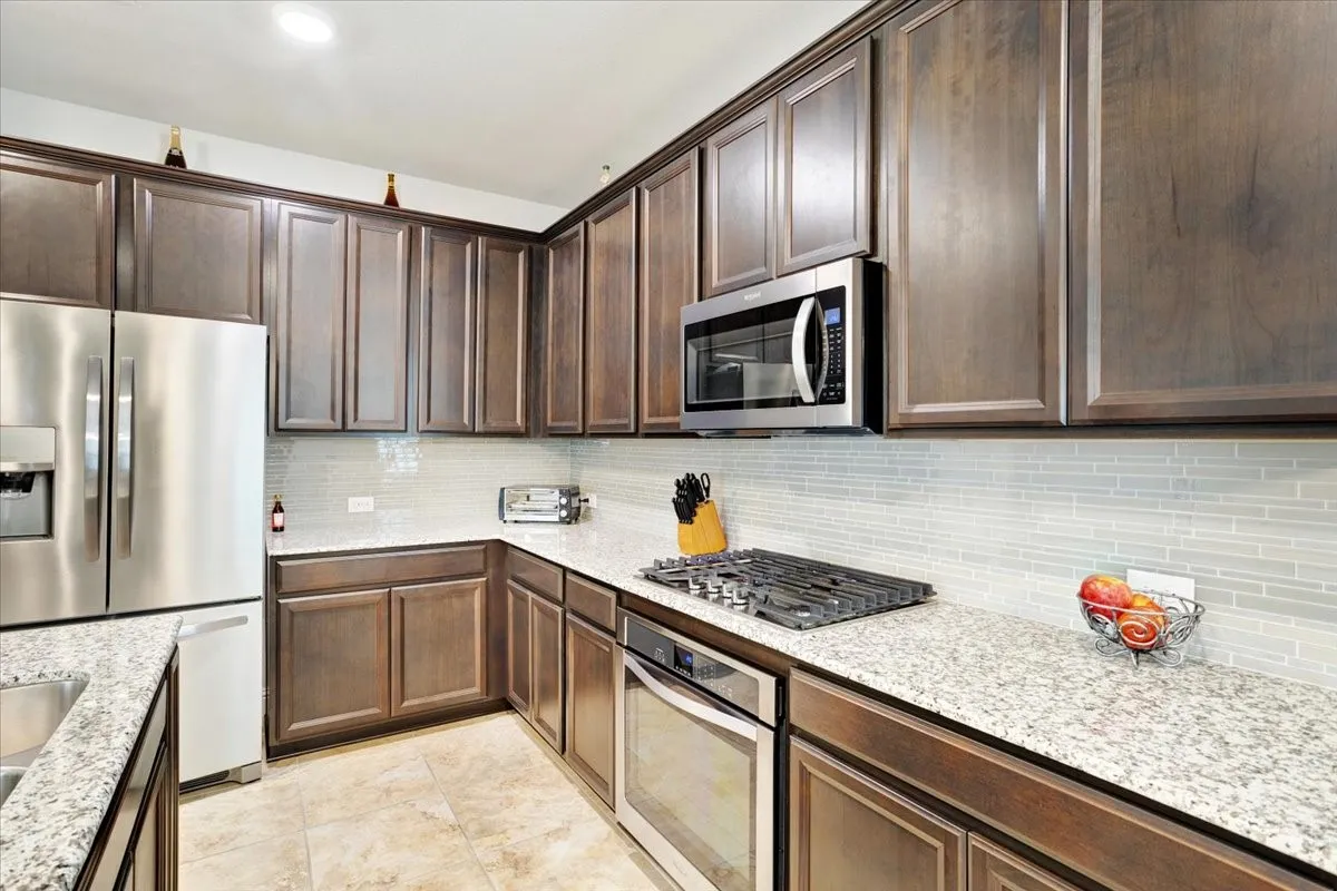 Elegant kitchen featuring rich wood-finish cabinetry with raised-panel doors, stainless steel appliances including a gas cooktop and built-in microwave, light-filled subway tile backsplash, and granite countertops throughout