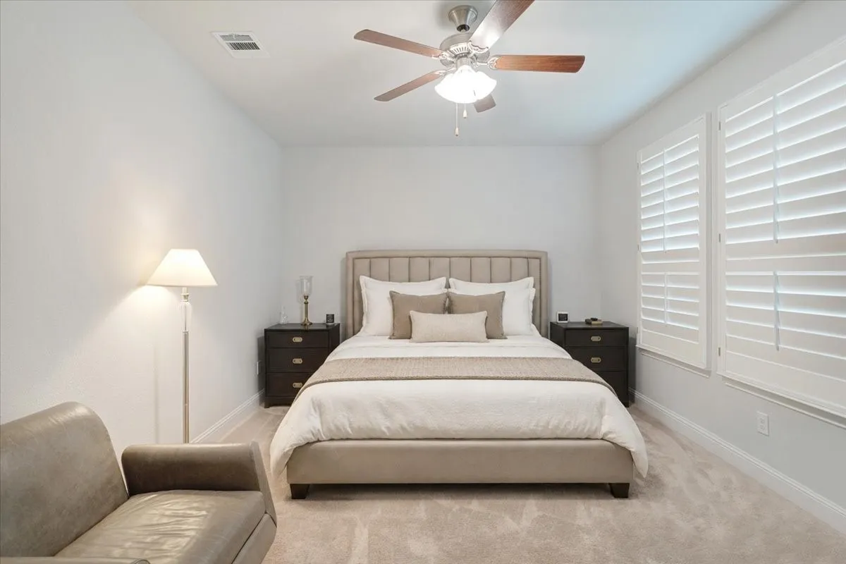 Neutral wall color, ceiling fan with light fixture, two windows with plantation shutters, light-colored carpet flooring, and white baseboards