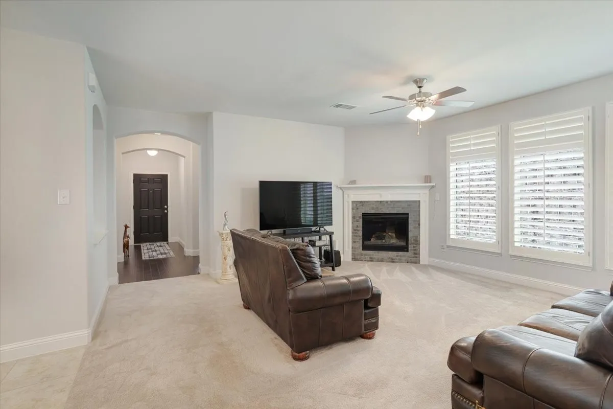 Carpeted living area featuring a fireplace with stone surround and white mantle, multiple windows with plantation shutters, and a ceiling fan with integrated lighting