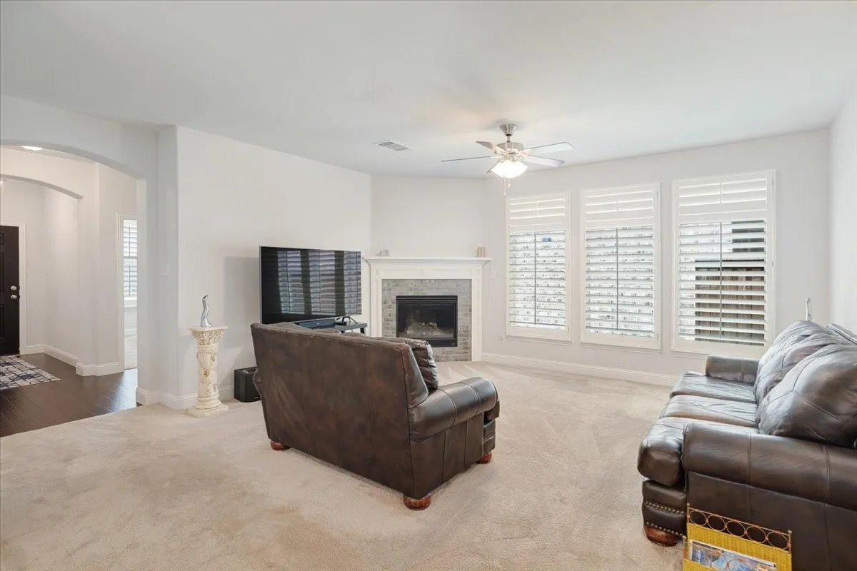 Carpeted living area featuring a central fireplace with a tile surround and mantel, three windows with white shutters, a ceiling fan, and an arched entry