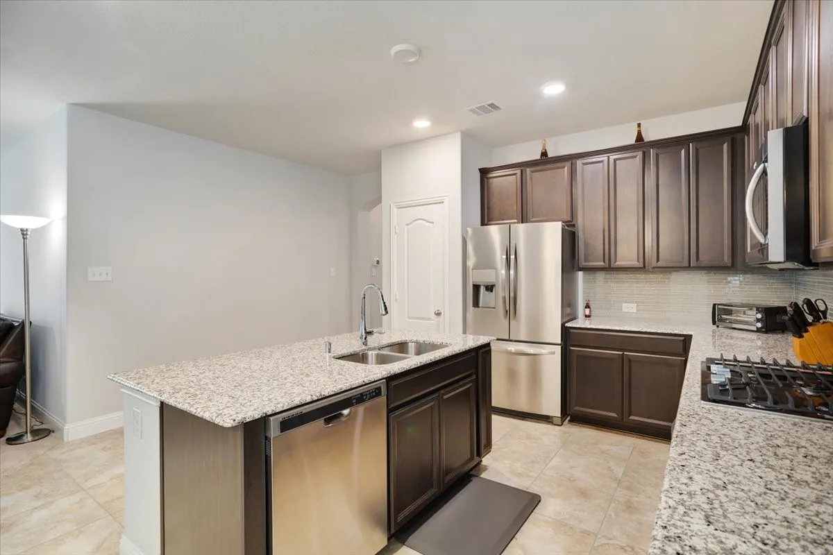 Modern kitchen featuring stainless steel appliances, granite-style countertops, dark wood cabinetry with brushed metal hardware, a center island with seating space, gas cooktop, and light wood-finish flooring throughout the open layout
