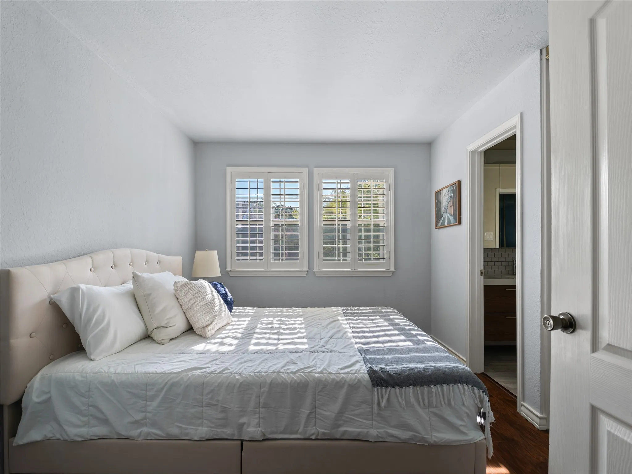 Two windows with white plantation shutters, an upholstered tufted headboard, and wood-finish flooring