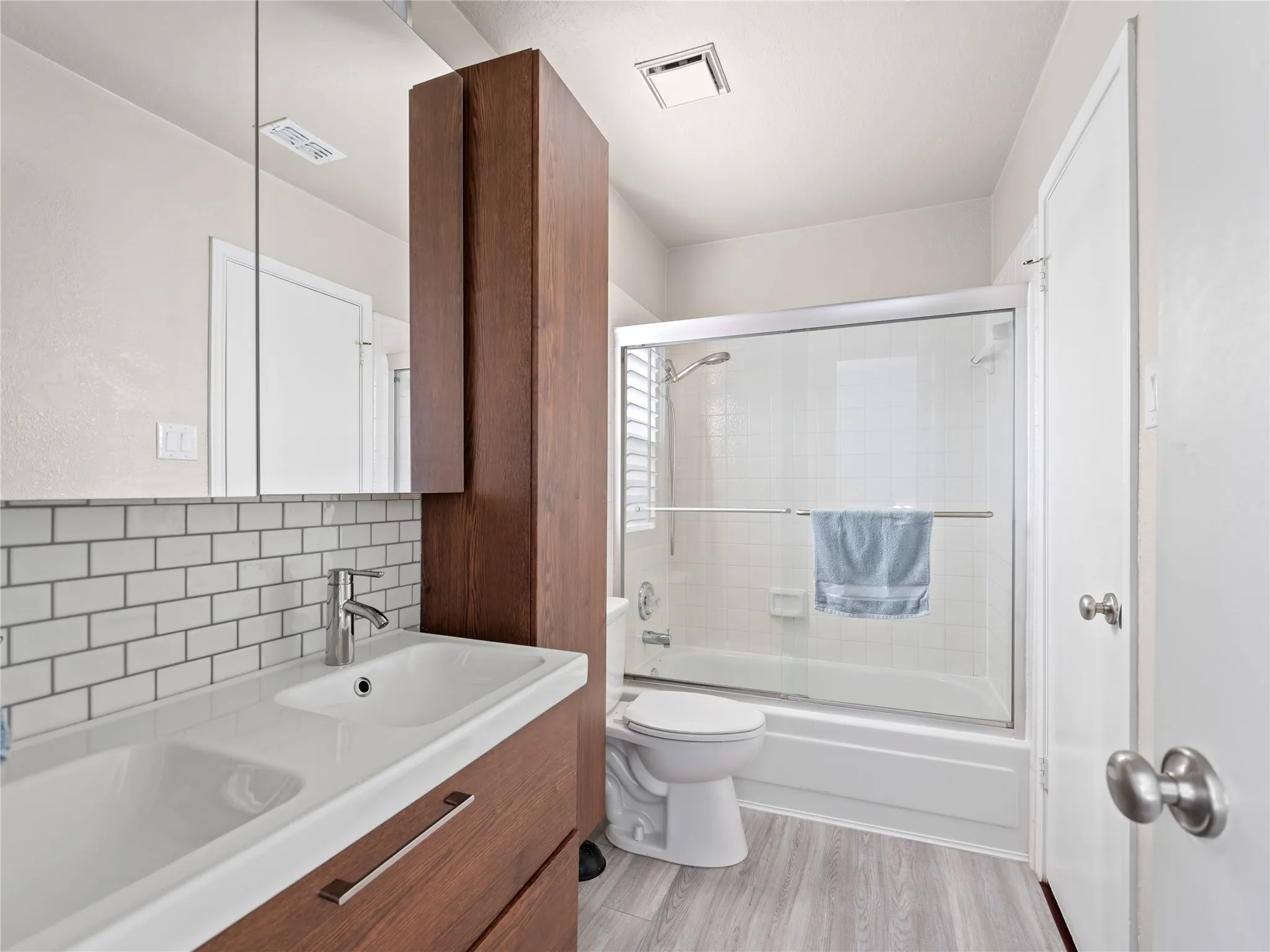 Vanity with integrated dual sinks, subway tile backsplash, wood-finish cabinetry, and a shower-tub combination with sliding glass doors