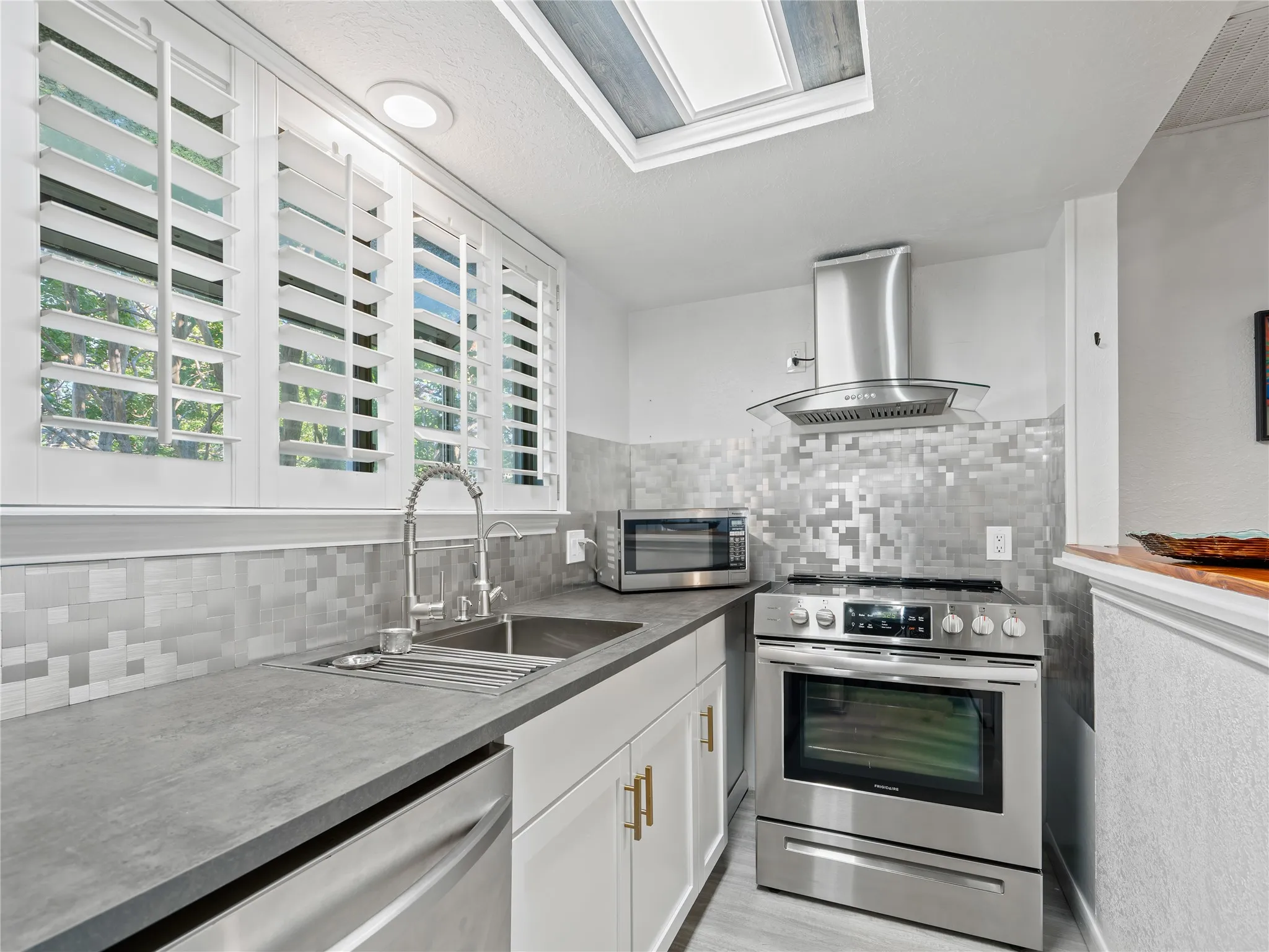 Kitchen featuring stainless steel appliances, solid surface countertops, a mosaic tile backsplash, a skylight, and white plantation shutters