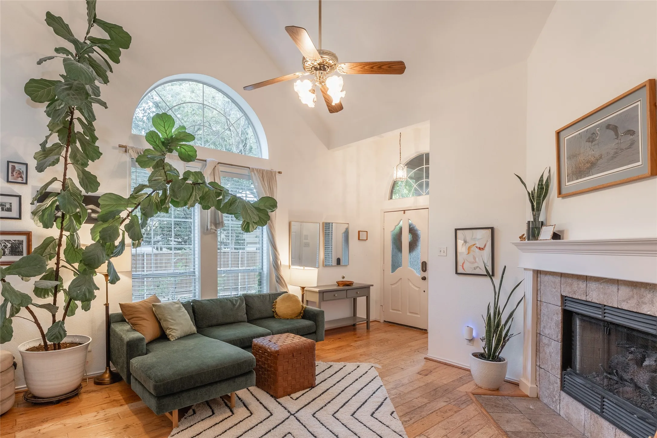 Vaulted ceiling with a ceiling fan, arched transom window, wood-finish flooring, tiled fireplace surround, and a white front door with an arched glass insert