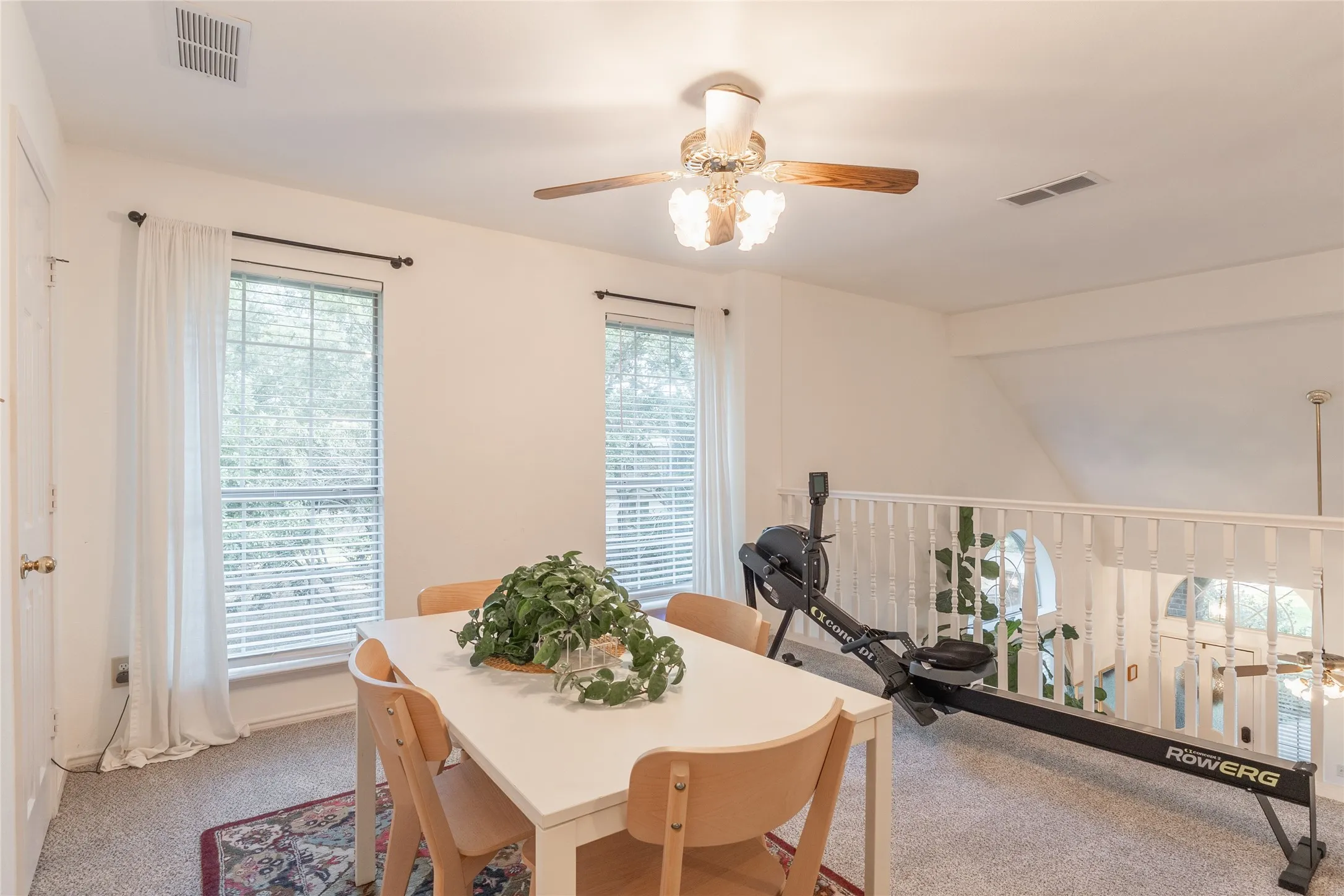 Loft space featuring two windows with blinds, a ceiling fan with light fixture, and a white baluster railing