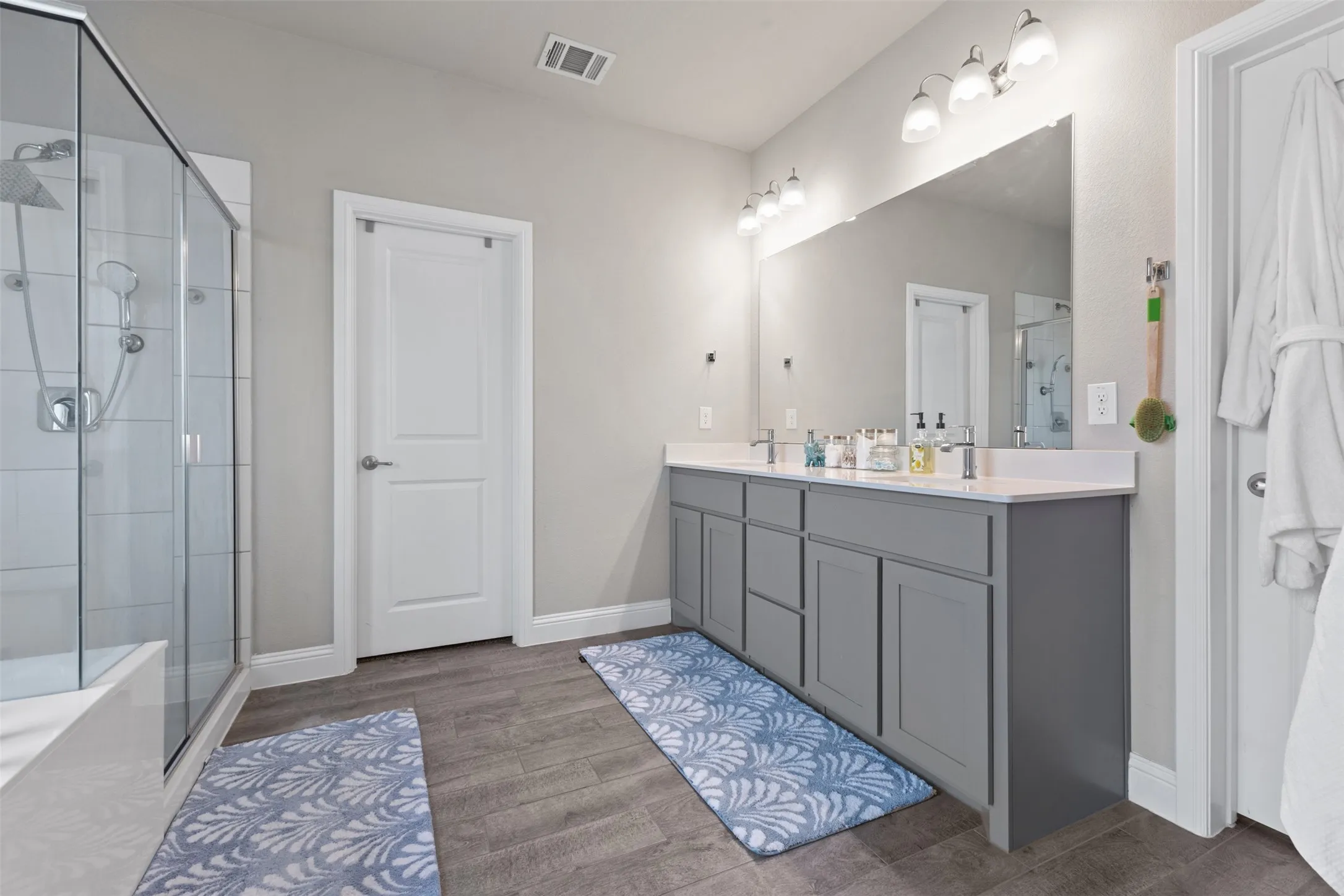 Dual vanity with gray cabinetry and white countertops, large wall-mounted mirror, modern light fixture, wood-finish flooring, and a glass-enclosed shower