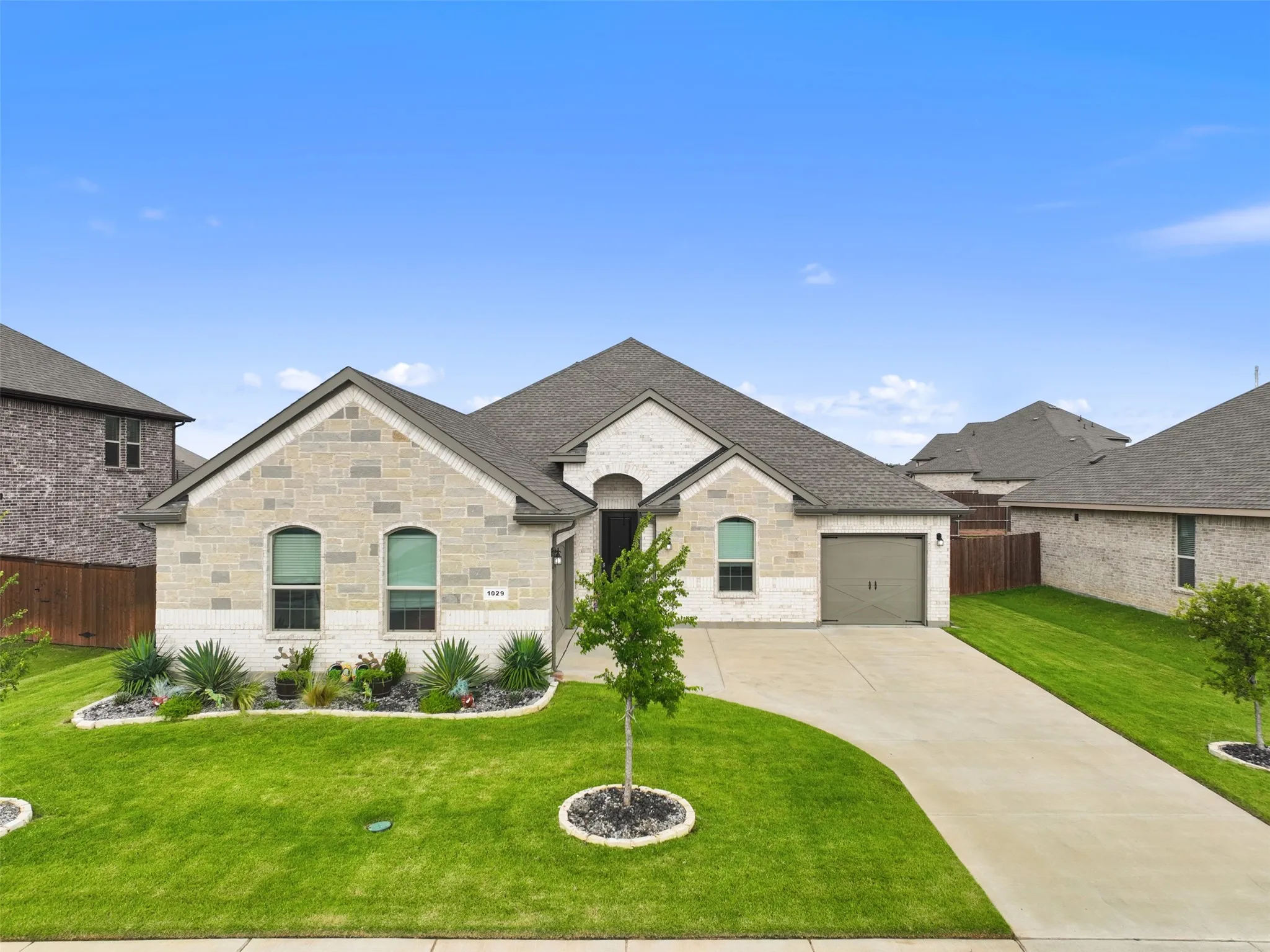 Stone and brick facade residence with an attached garage, featuring a concrete driveway, manicured lawn, and landscaped front beds